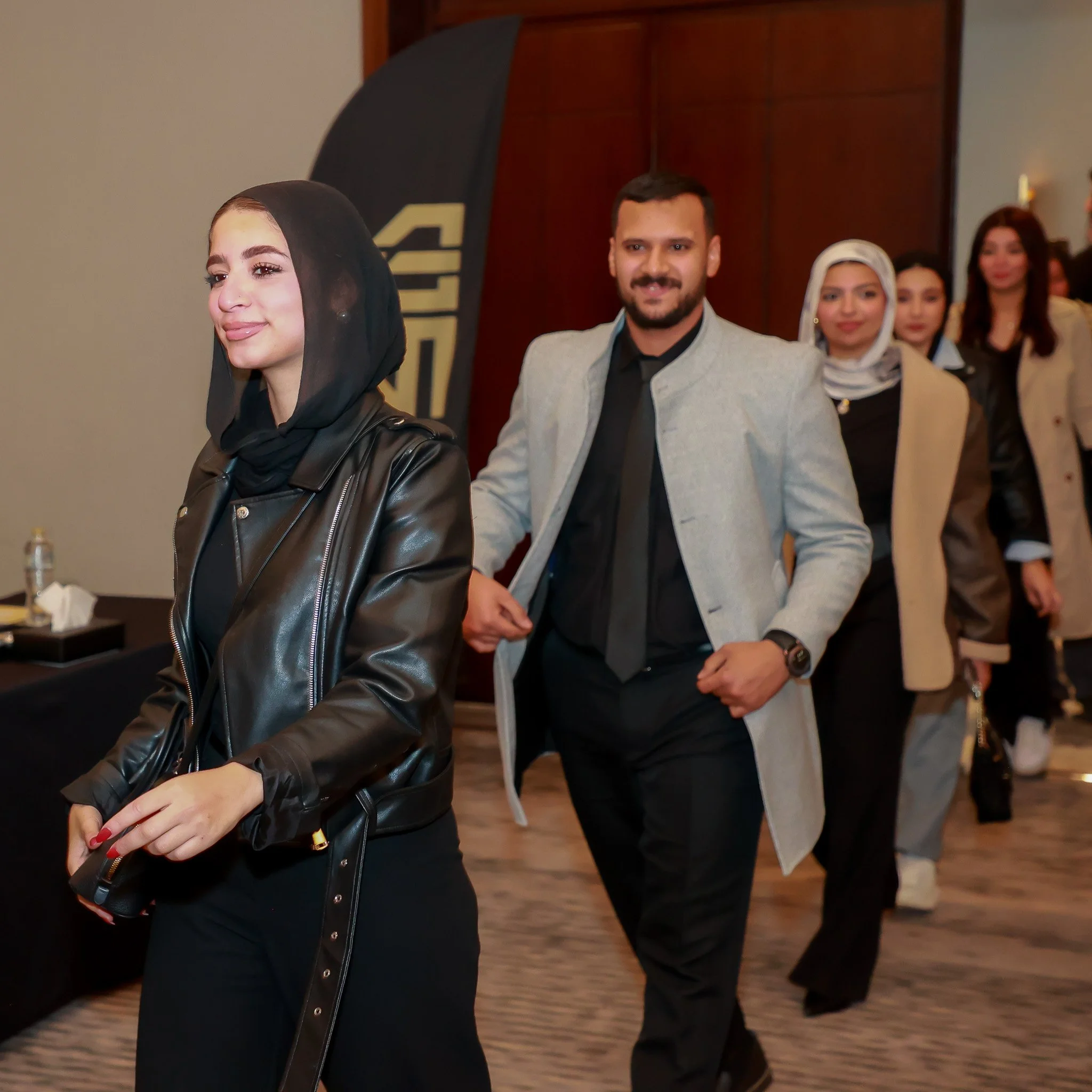 A group of diverse young adults walking in line at an indoor event, with a woman in a black headscarf and leather jacket leading, followed by a man in a gray blazer and black tie, and others in stylish attire.