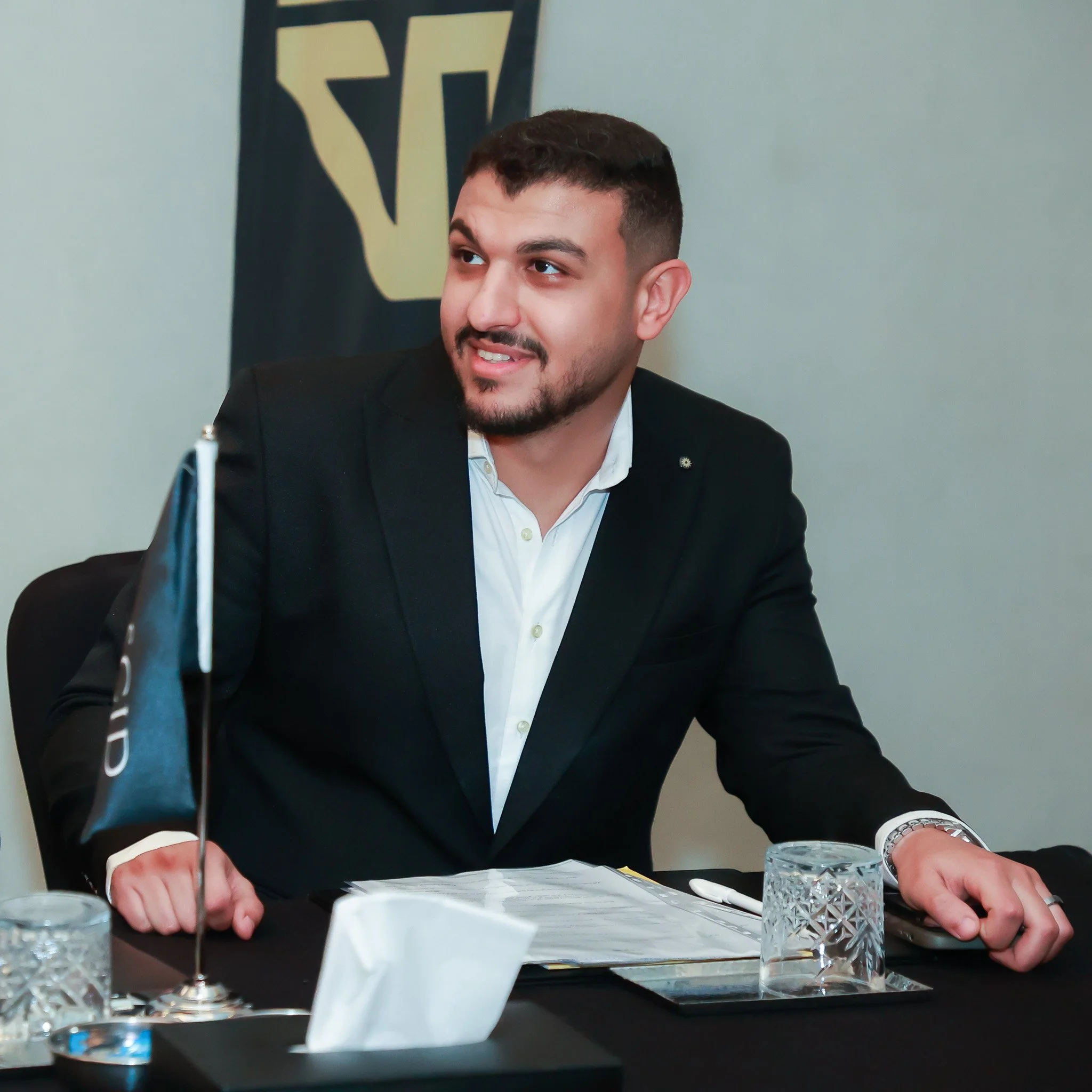 A man in a black suit and white shirt sitting at a conference table with documents, a pen, glasses, a flag, and a tissue box, in front of a USA flag backdrop.
