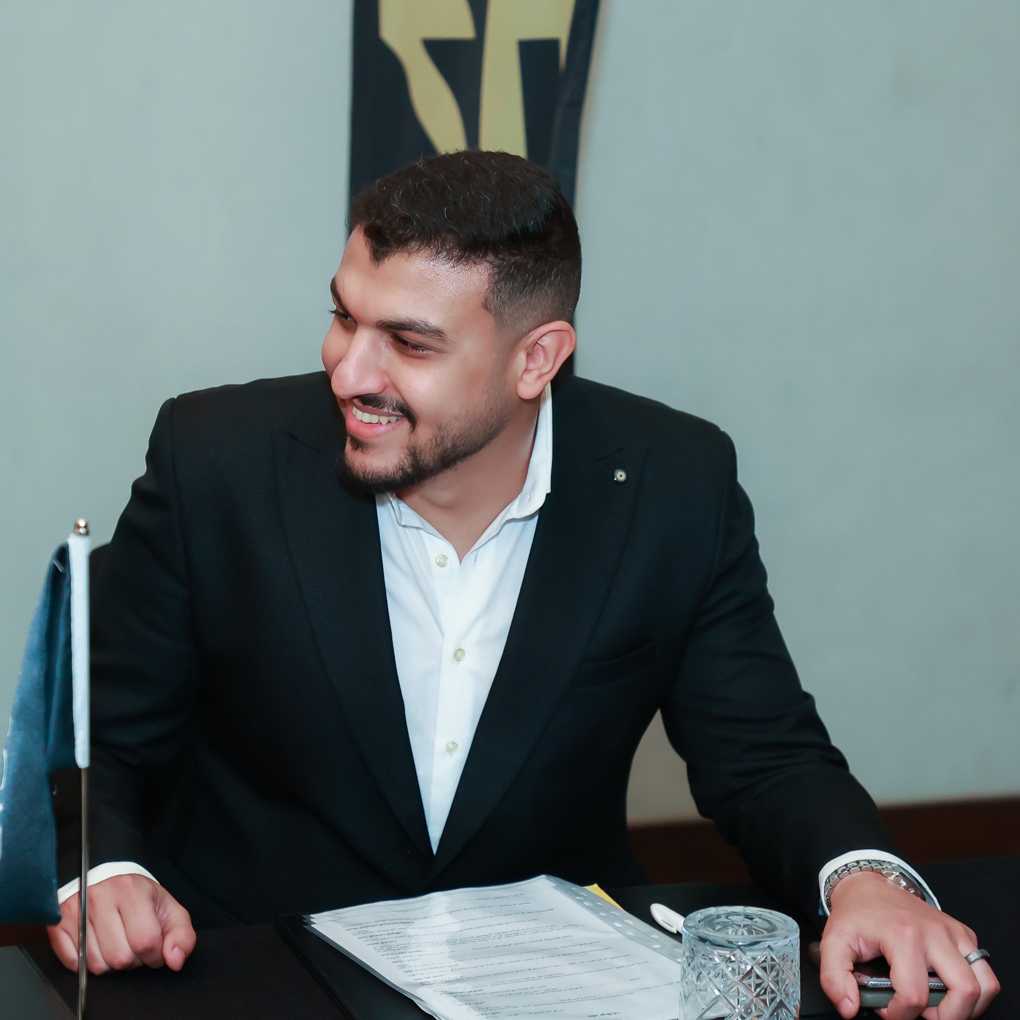 A smiling man in a black suit and white shirt sits at a desk with papers, a pen, and a glass of water, in a professional setting.