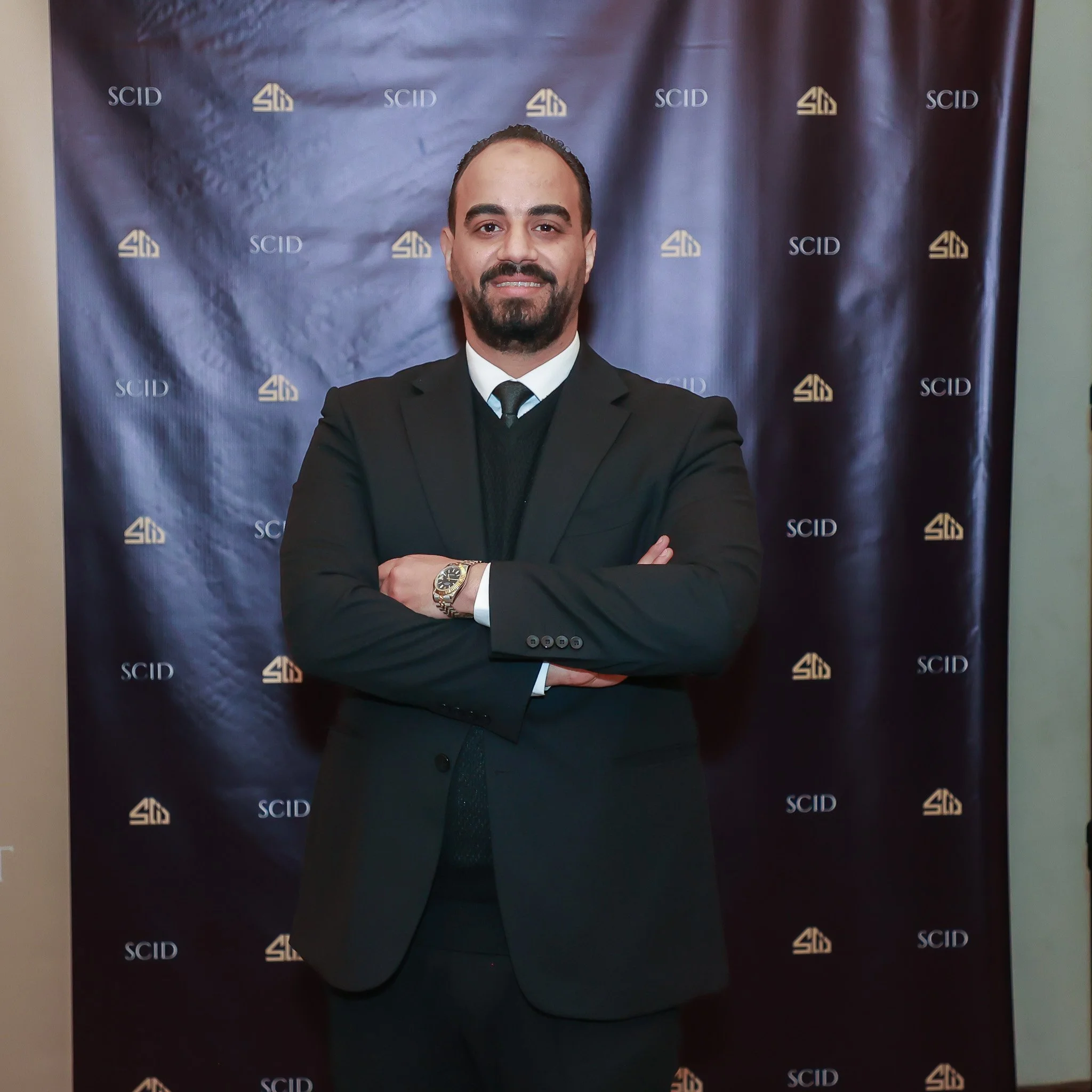 A man in a black suit, white shirt, and black tie standing with arms crossed in front of a blue backdrop featuring the logos 'SCID' and a geometric shield symbol.