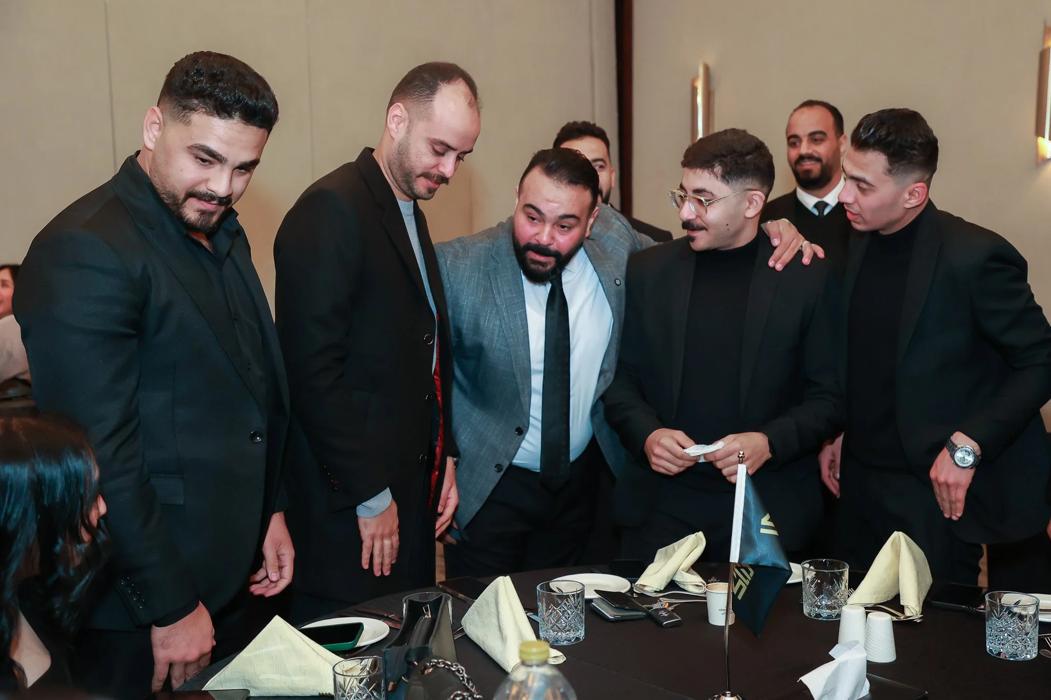 Group of six men in suits gathered around a table with napkins and glasses, in a formal setting.