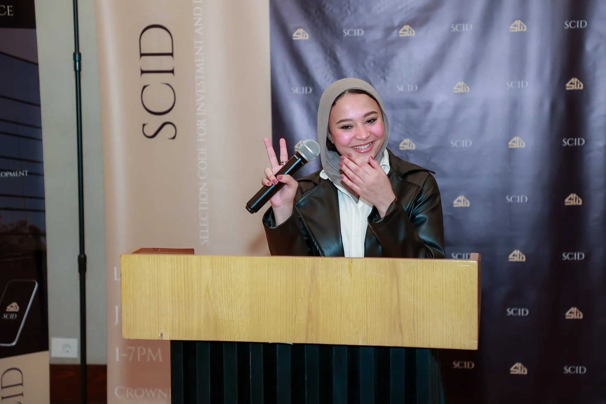 A young woman wearing a beige hijab and a black leather jacket standing behind a wooden podium, smiling and holding a microphone in her right hand, making a peace sign with her left hand, in front of a backdrop with the SCID logo.