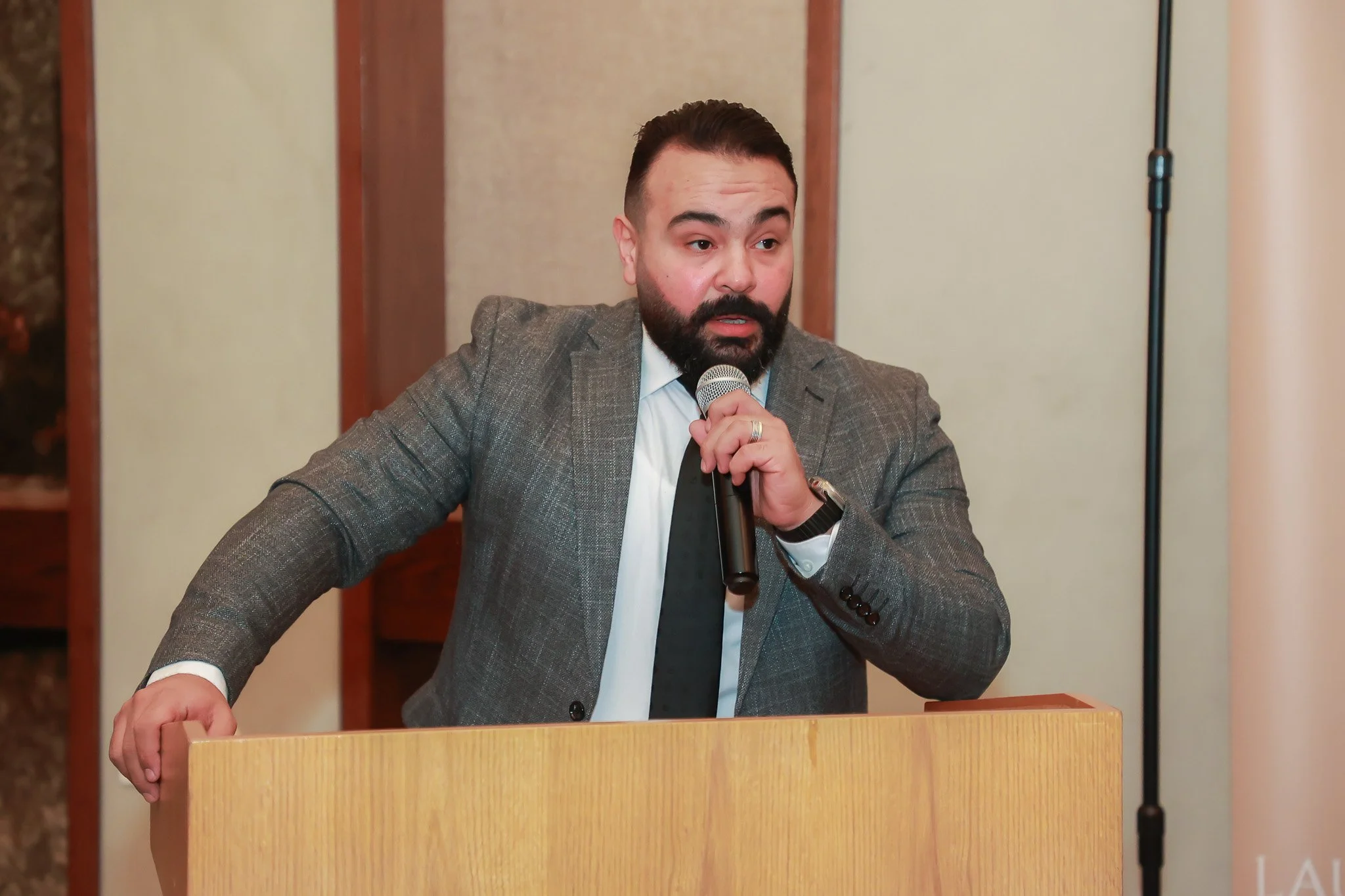 A man with a beard and short dark hair, in a gray suit, black tie, and white shirt, speaking into a microphone at a wooden podium.
