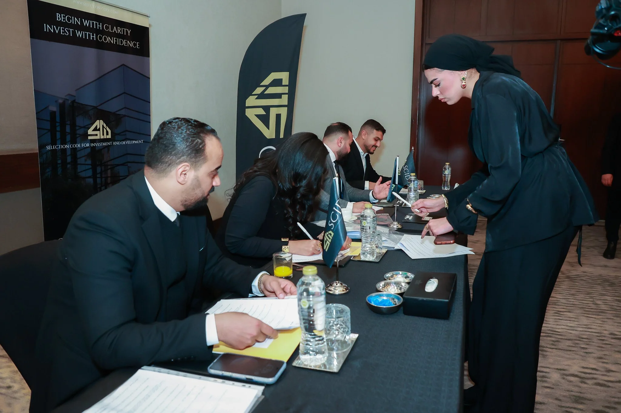 People sitting at a table during a conference or meeting, with informational banners and flags, and a woman standing and speaking to the seated individuals.