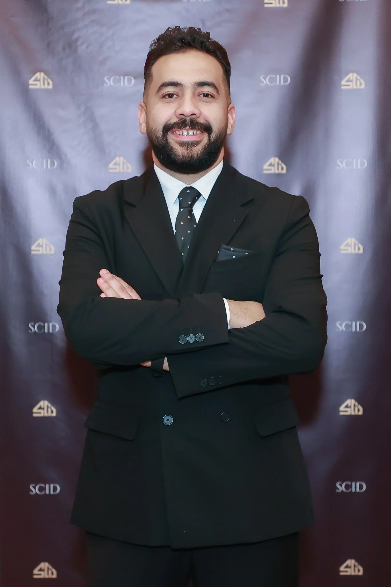 A man in a black suit with a white shirt and a black polka dot tie, standing with arms crossed and smiling, against a backdrop with the logo and text 'SCID' and '4D' in gold and white.