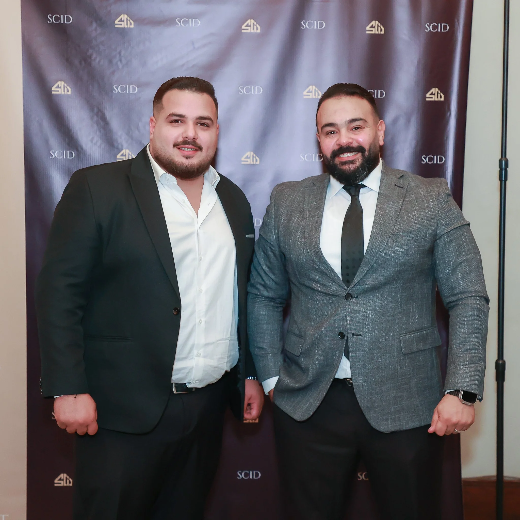 Two men in suits smiling and standing together in front of a backdrop with the logo 'SCID' repeated across it.