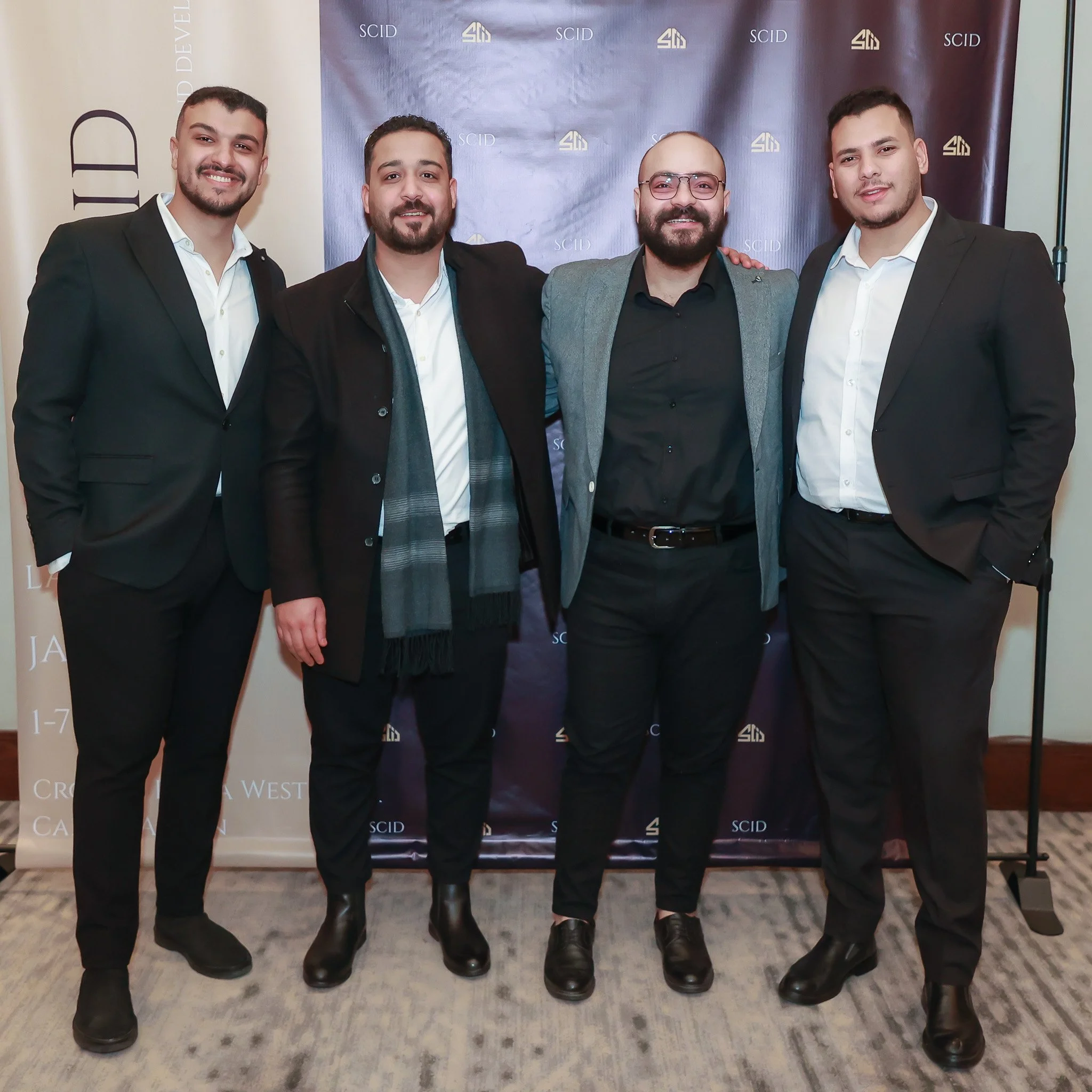 Four men in formal attire standing together indoors, posing for a photo in front of a backdrop with logos and text.