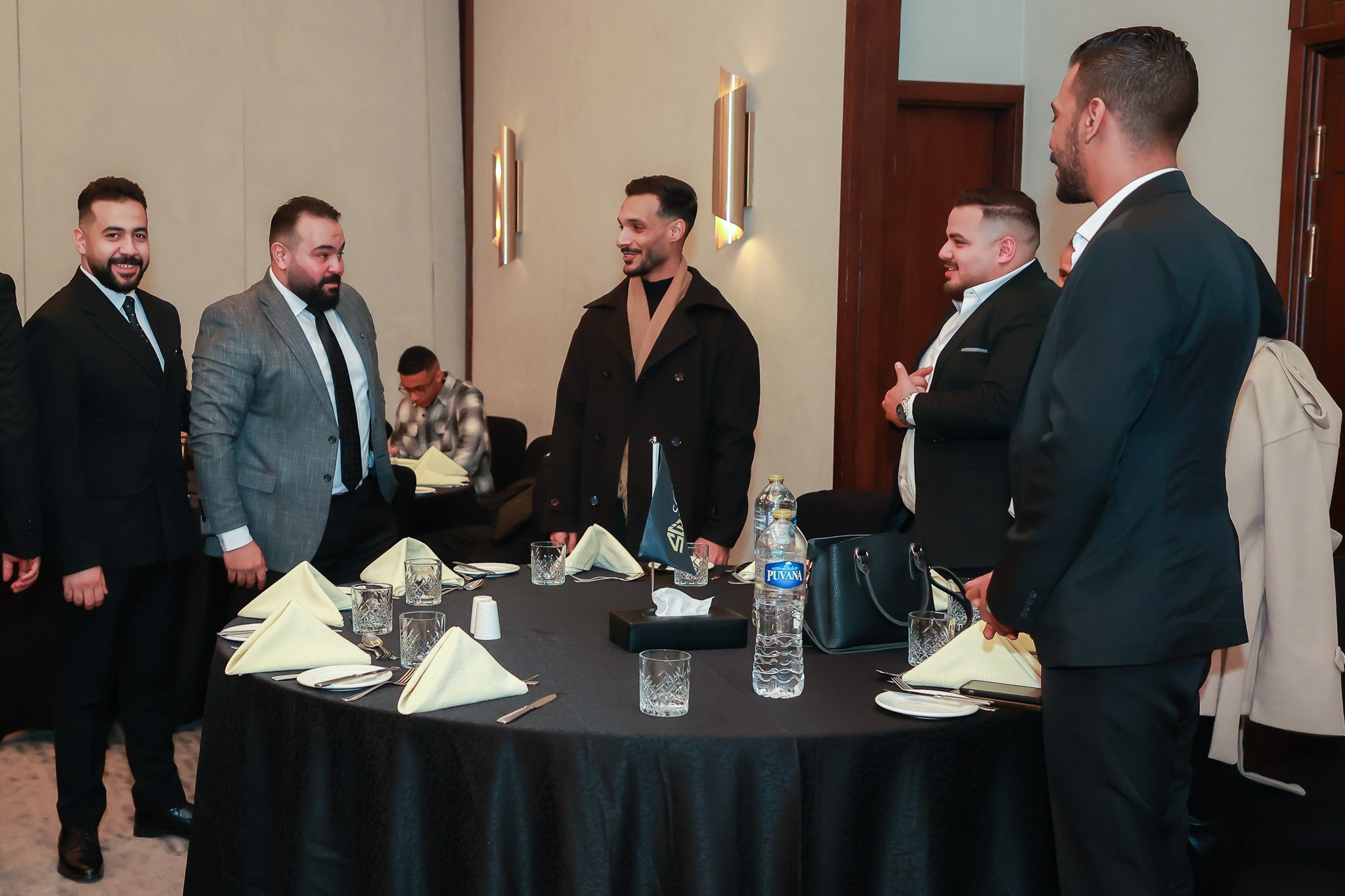 Group of six men in suits standing around a table set for a meal, engaged in conversation in a formal dining room.