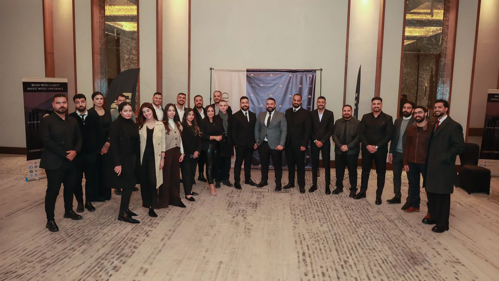 Group of professionally dressed people standing together at an indoor event with banners and a backdrop behind them.