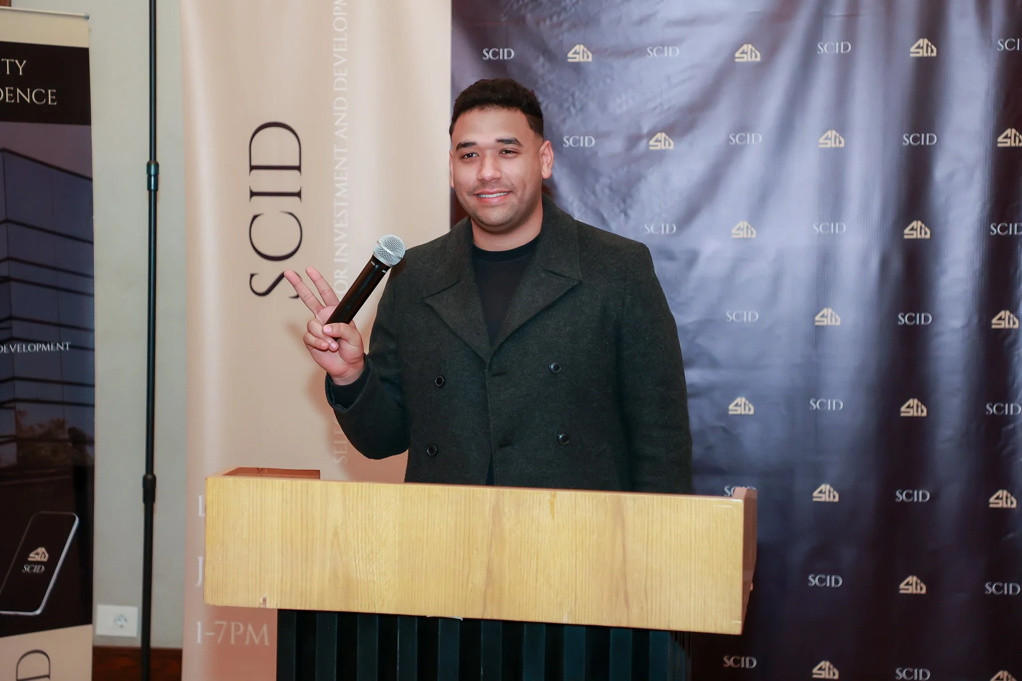 A man smiling and holding up a peace sign with his right hand at a podium with a microphone. Behind him are banners that say SCI Development and Silicon Valley Investment & Development.