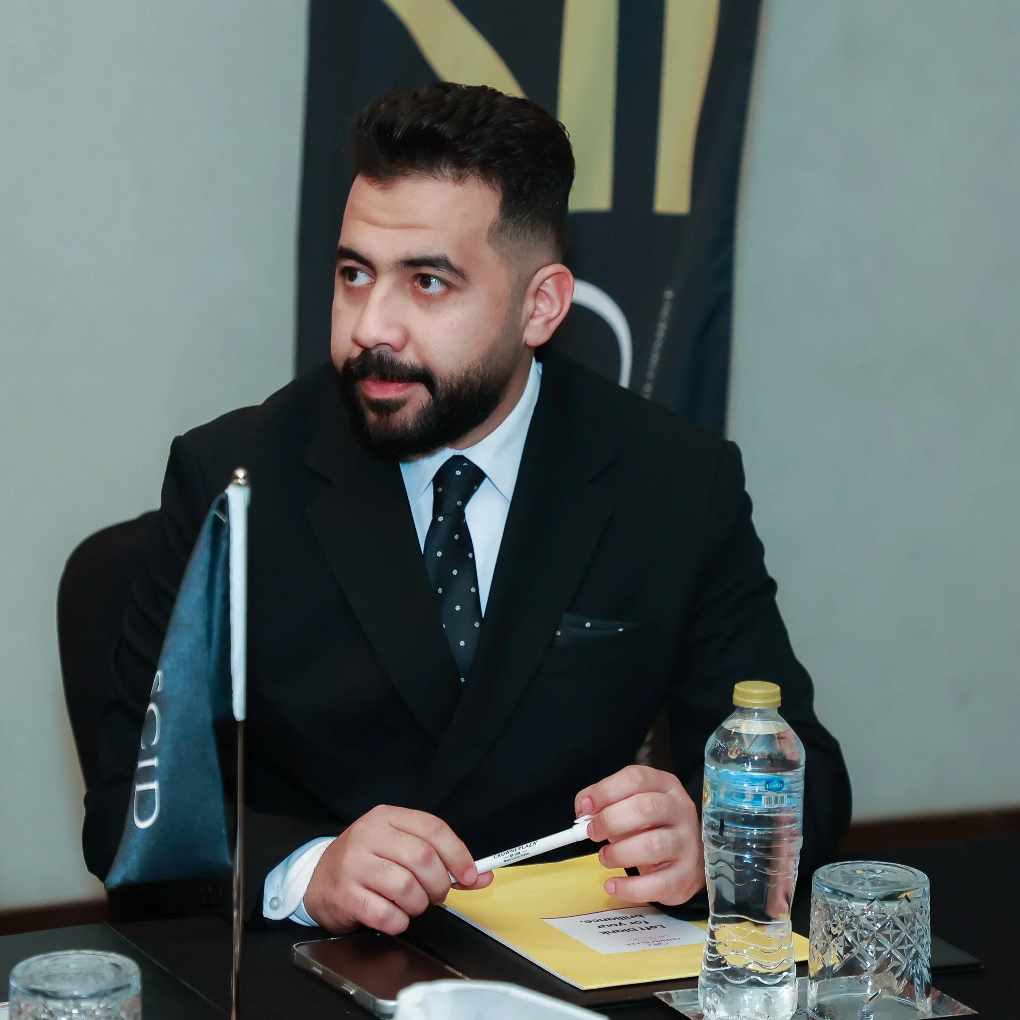 A man in a black suit and tie sitting at a conference table with a flag, water bottle, glass, and yellow folder, engaged in a meeting or discussion.