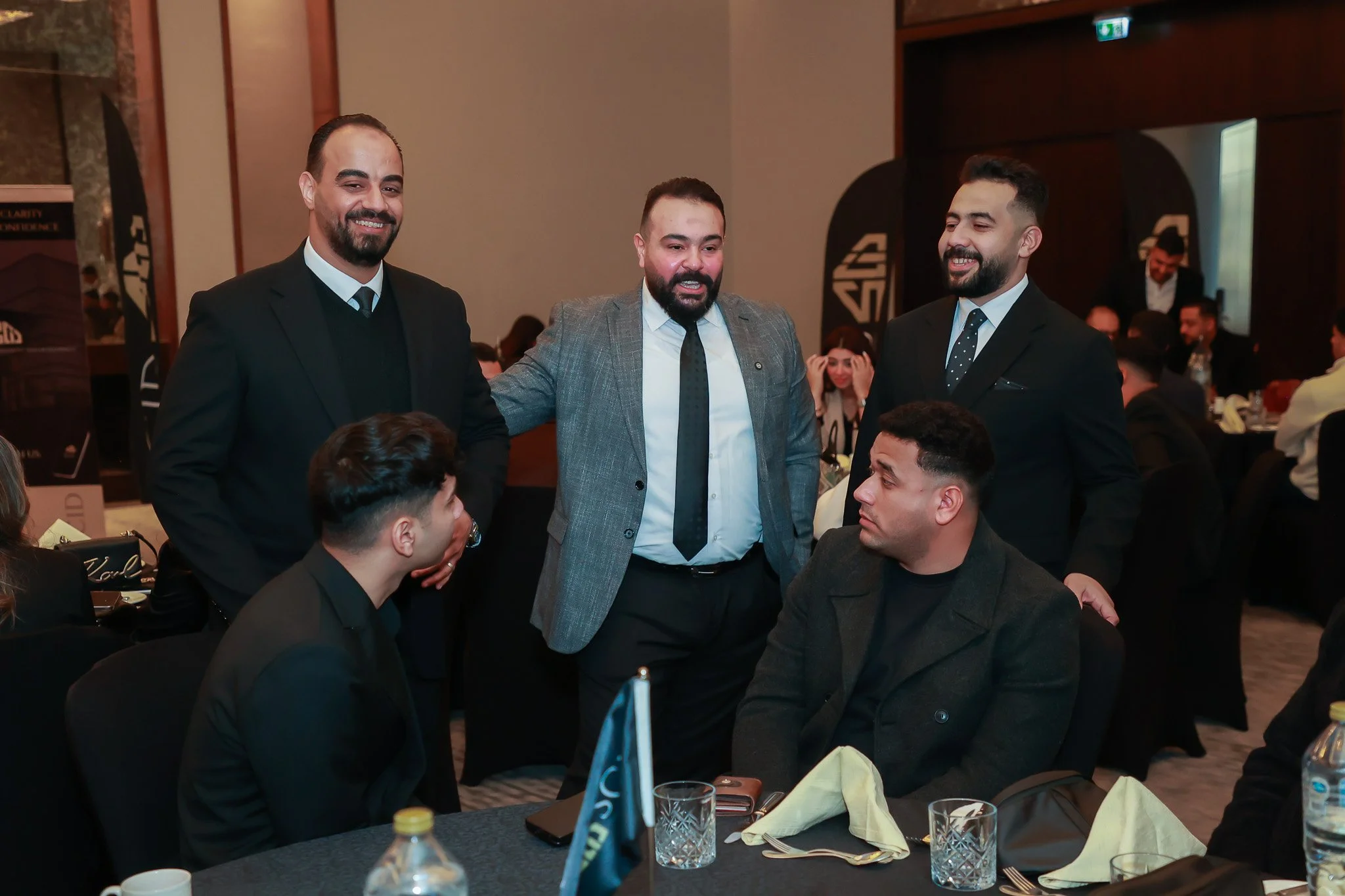 Five men in formal attire at a banquet table, engaged in conversation, with banners and other guests in the background.