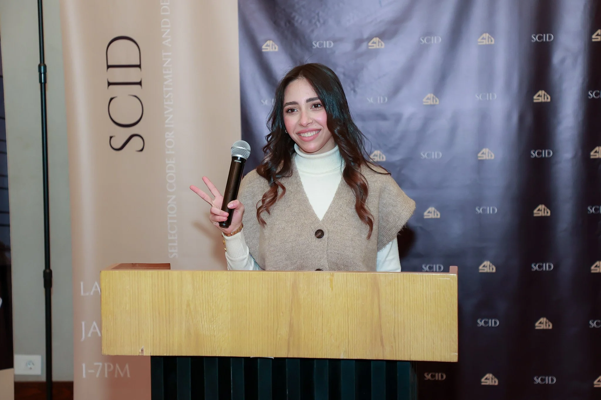 A young woman with long, wavy dark hair is smiling and holding a microphone with her right hand. She is standing behind a wooden podium, wearing a beige vest over a white turtleneck. The background has a navy blue banner with the SCID logo and a beig