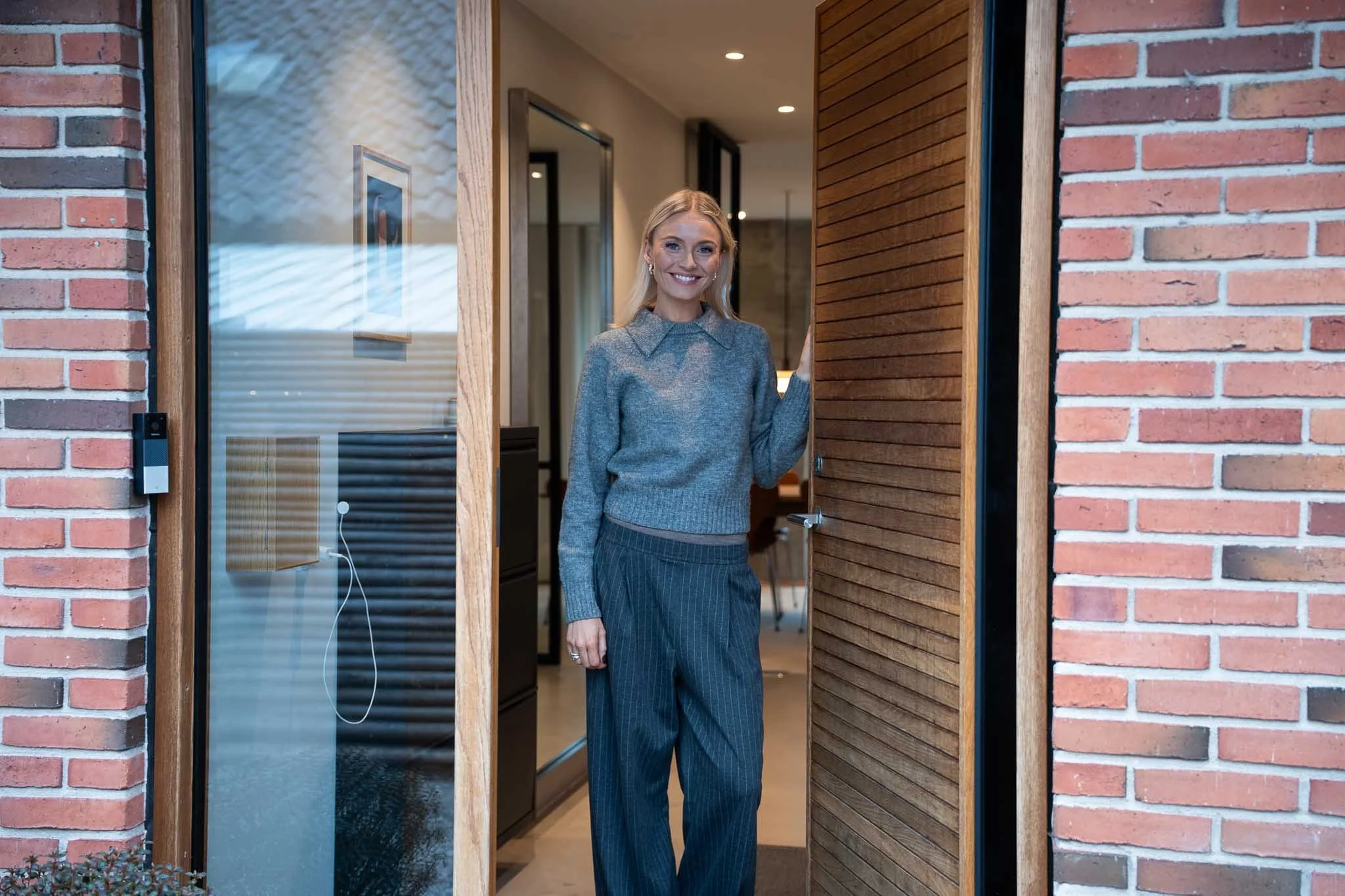 A woman with blonde hair wearing a gray sweater and pinstripe wide-leg pants, smiling and standing in the doorway of a house.