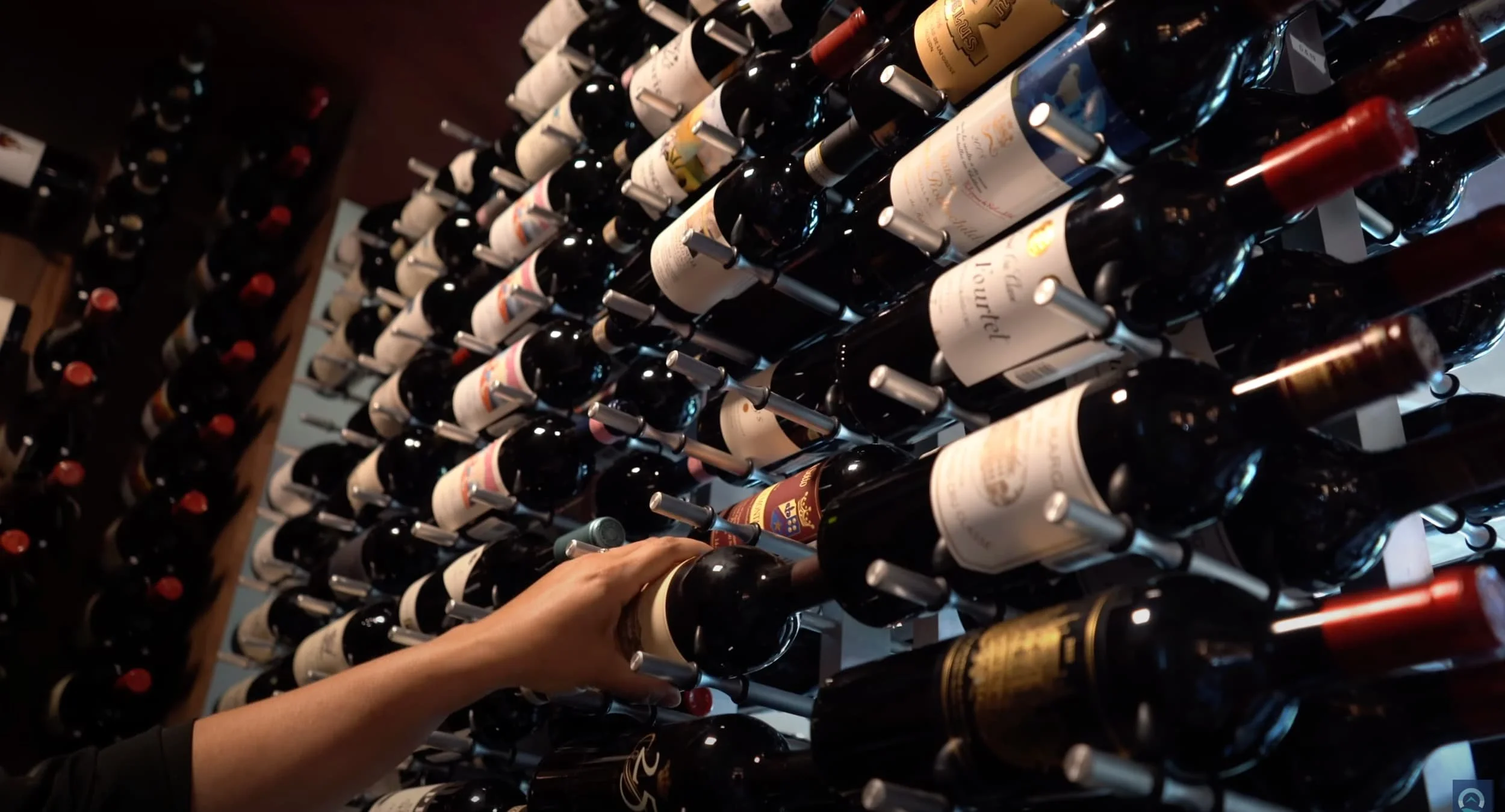 A person selecting a bottle of red wine from a rack filled with wine bottles in a wine cellar.