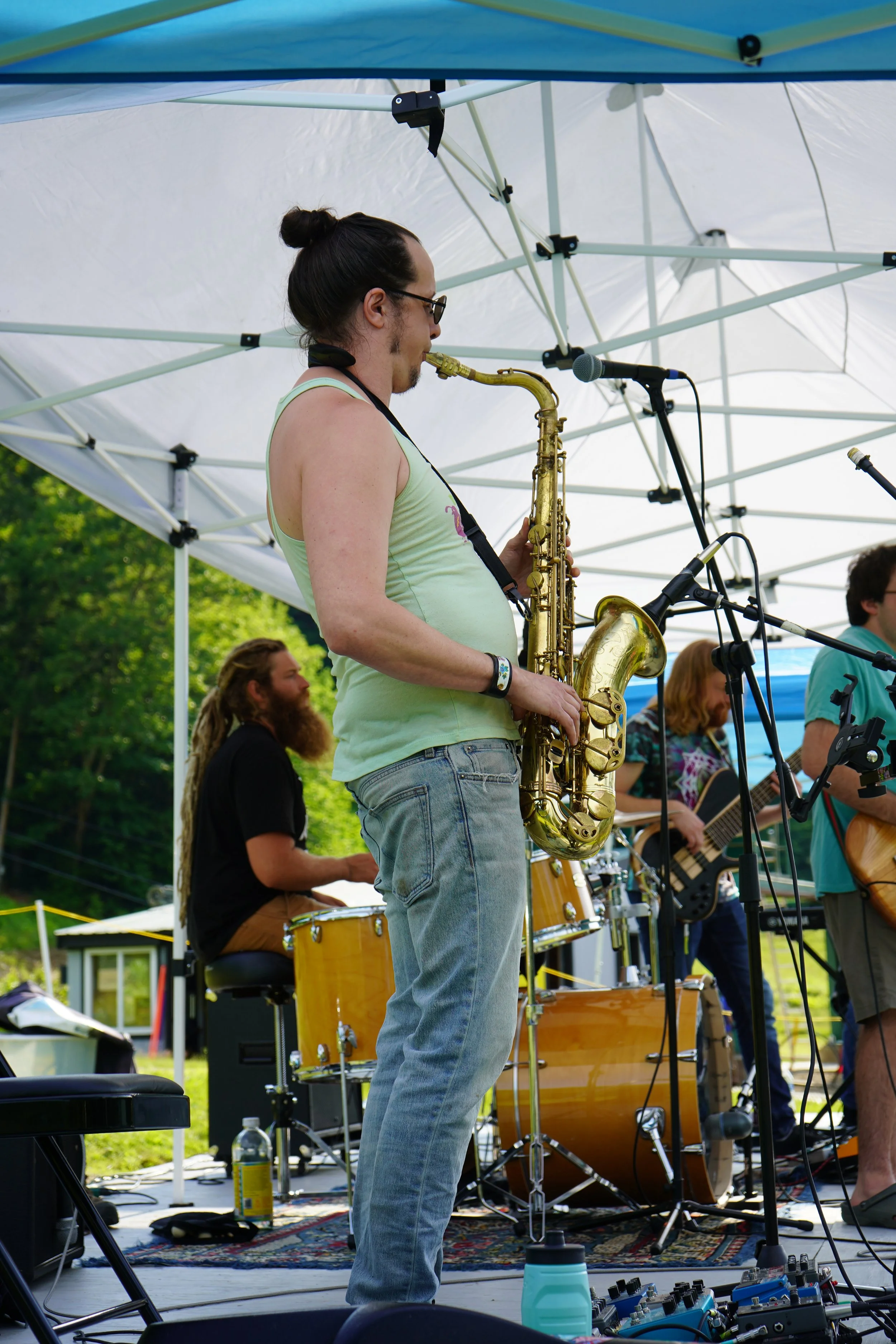 Musician playing saxophone on stage at outdoor music event with a drummer, guitarist, and vocals, under a white canopy with lush green trees in the background.