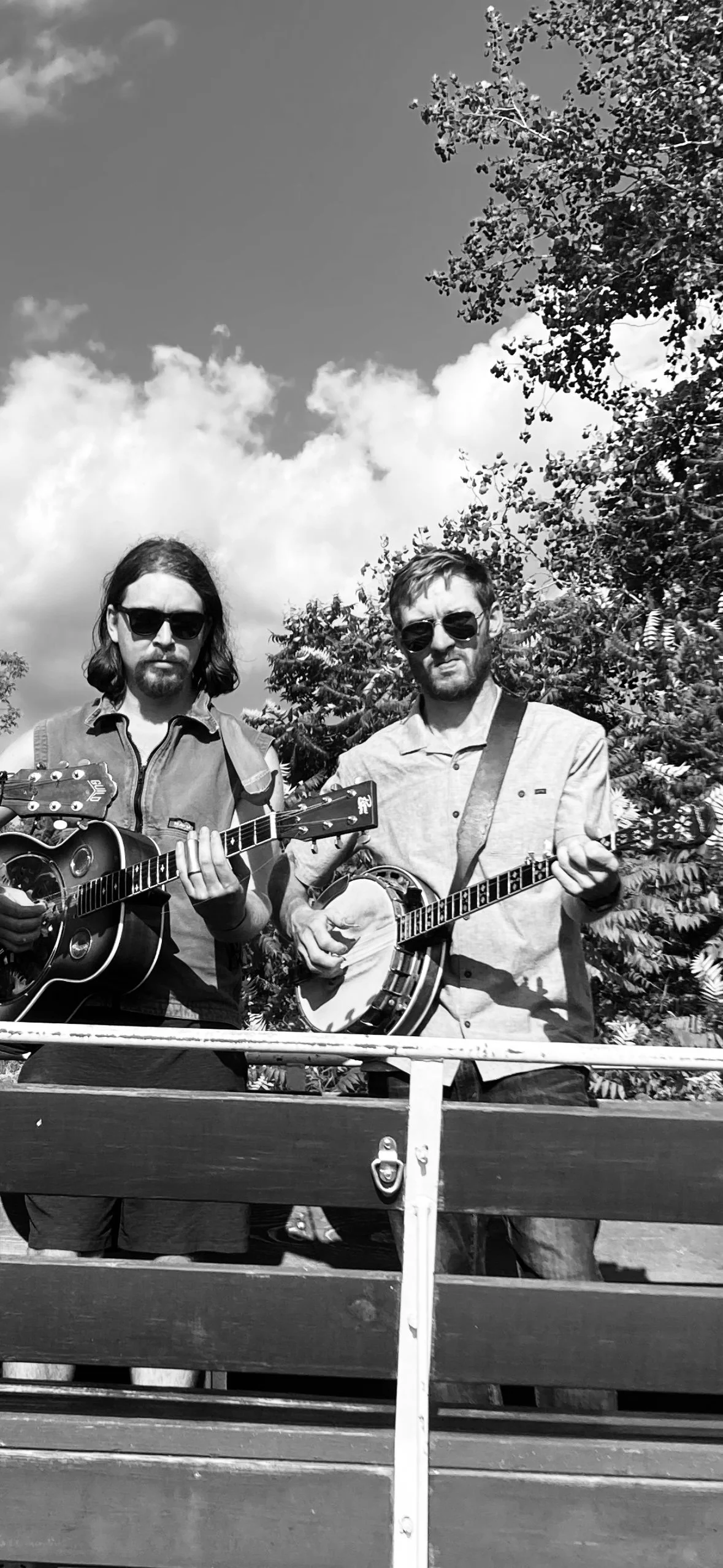 Two men wearing sunglasses, playing guitars and a banjo outdoors on a sunny day with trees and a cloudy sky in the background.