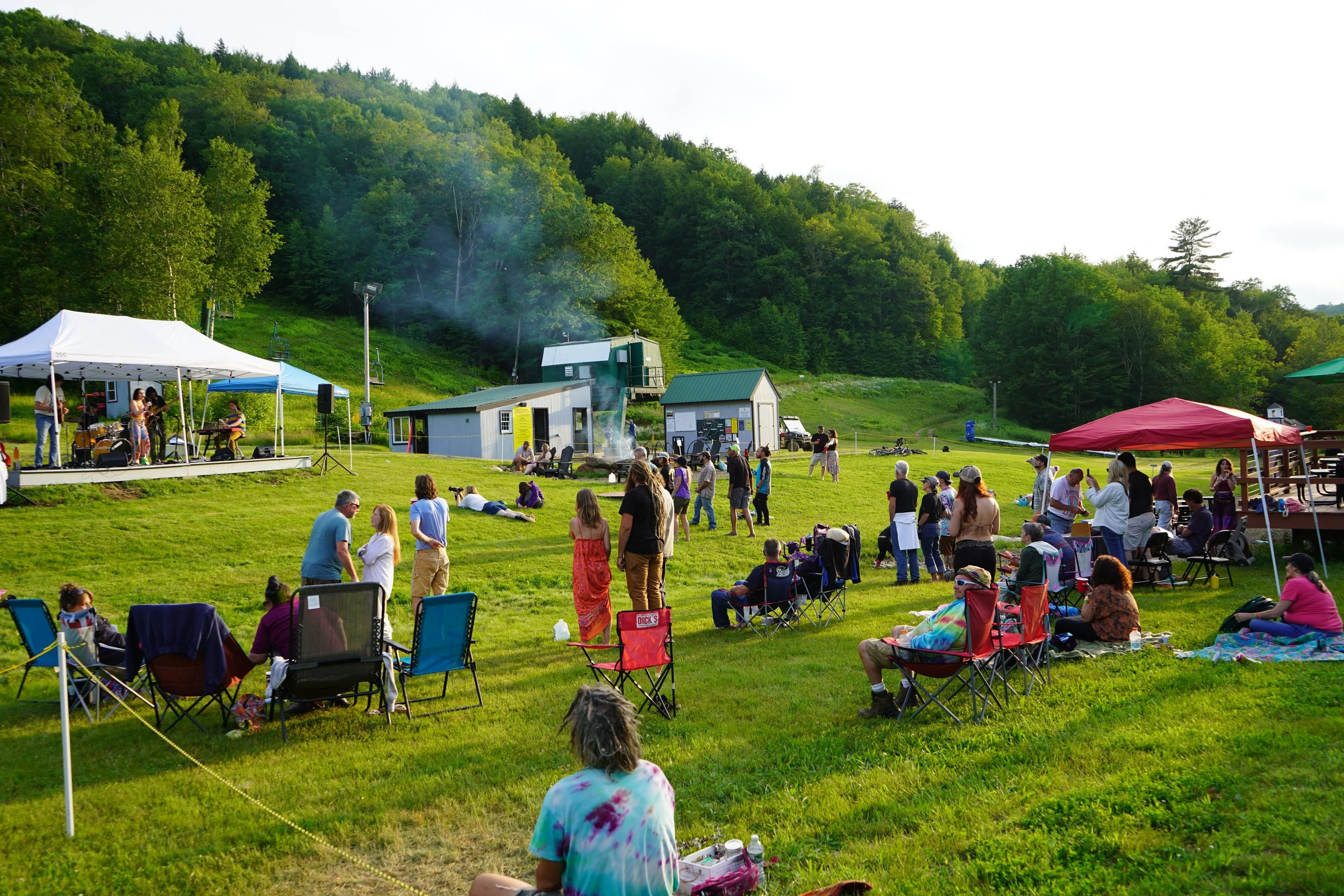 People gathered on a grassy hillside for an outdoor event with a small stage and live band, tents, and a lush green background of trees and hills.