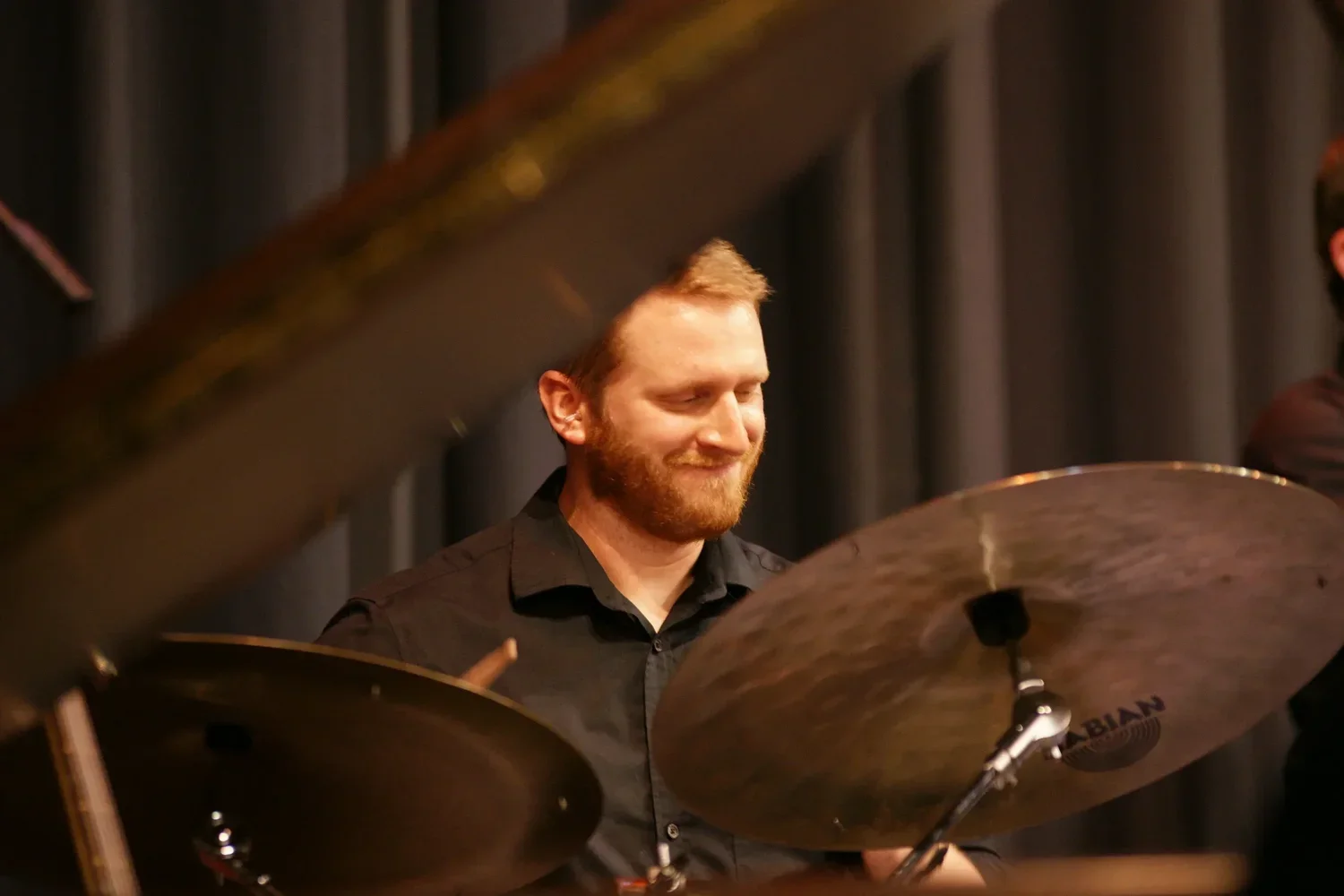 A man with a beard and short, light hair playing a cymbal and smiling, with dark curtains in the background.