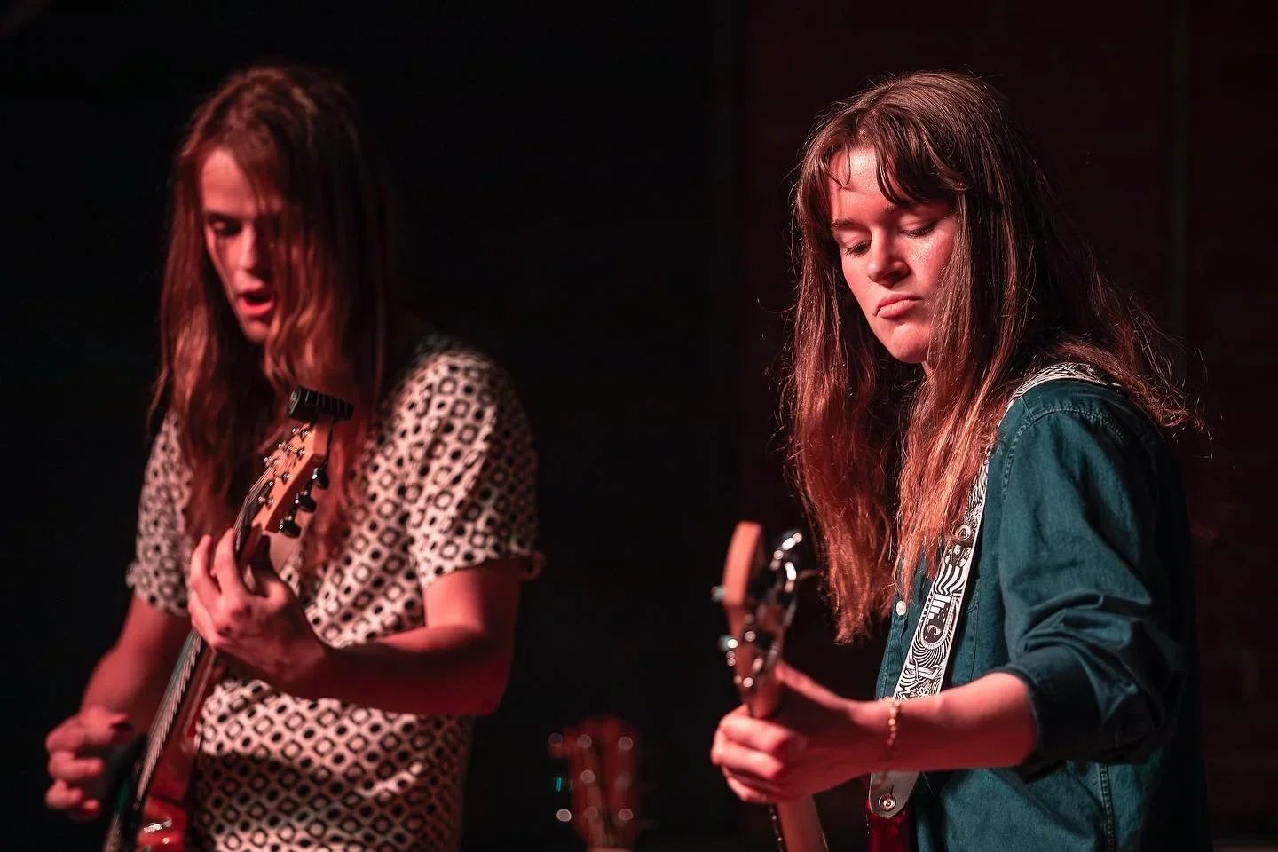 Two women with long hair playing guitars on stage in a dark venue.