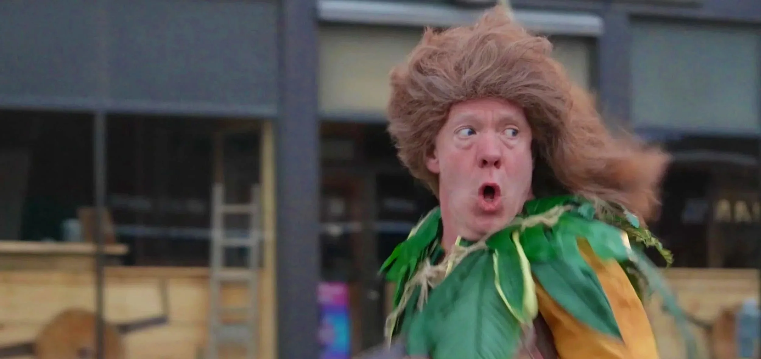 A man with long, wild hair and an surprised expression on his face, wearing a costume with large green leaves and holding a book