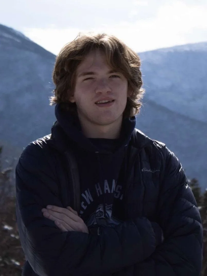 A young man with light brown, wavy hair and fair skin standing outdoors with his arms crossed, wearing a black jacket and hoodie, in front of a mountainous landscape with cloudy skies.
