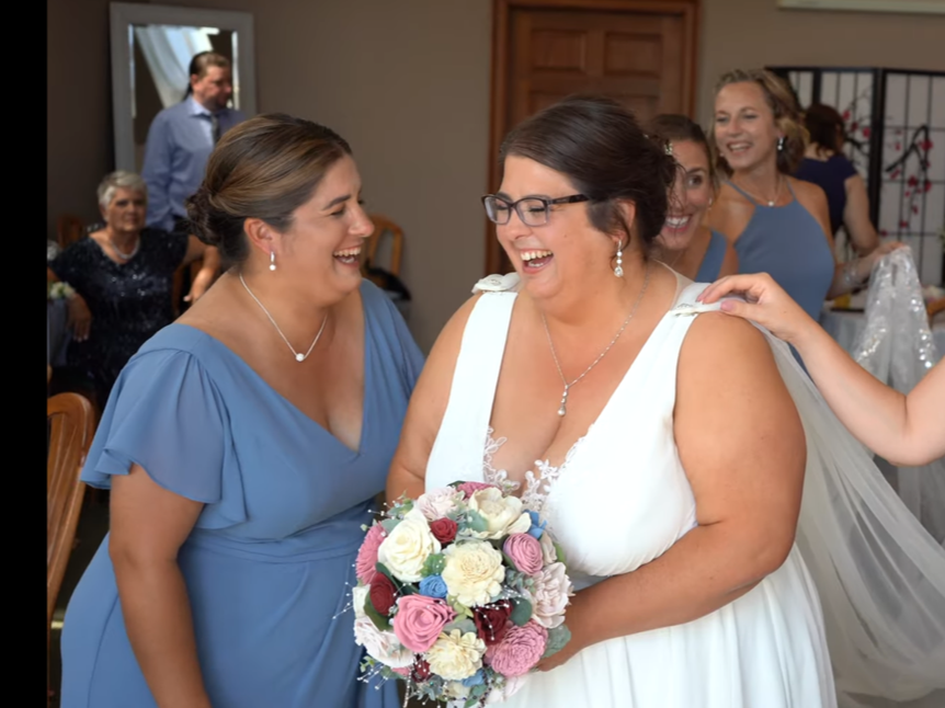 A bride in a white wedding dress holding a colorful bouquet, smiling and sharing a joyful moment with a woman in a blue dress at a wedding celebration, with other guests in the background.