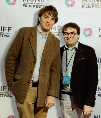 Two young men standing together at a film festival, one in a brown blazer and the other in a dark blazer, both smiling for the photo. Behind them is a backdrop with event logos.