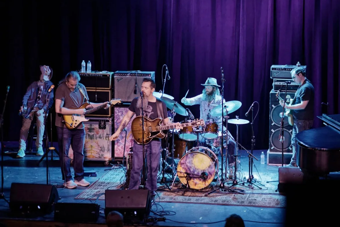 A band performing on stage with five members playing guitars, drums, and keyboard, with purple curtains in the background.