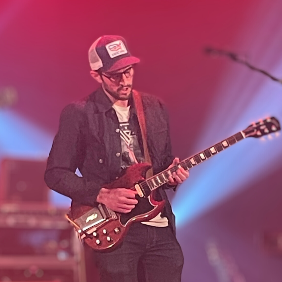 A man playing an electric guitar on stage with colorful lighting in the background.