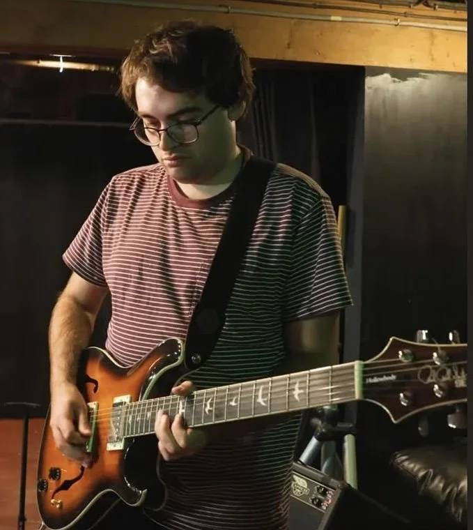 A young man with glasses and dark hair playing an electric guitar in a music studio.