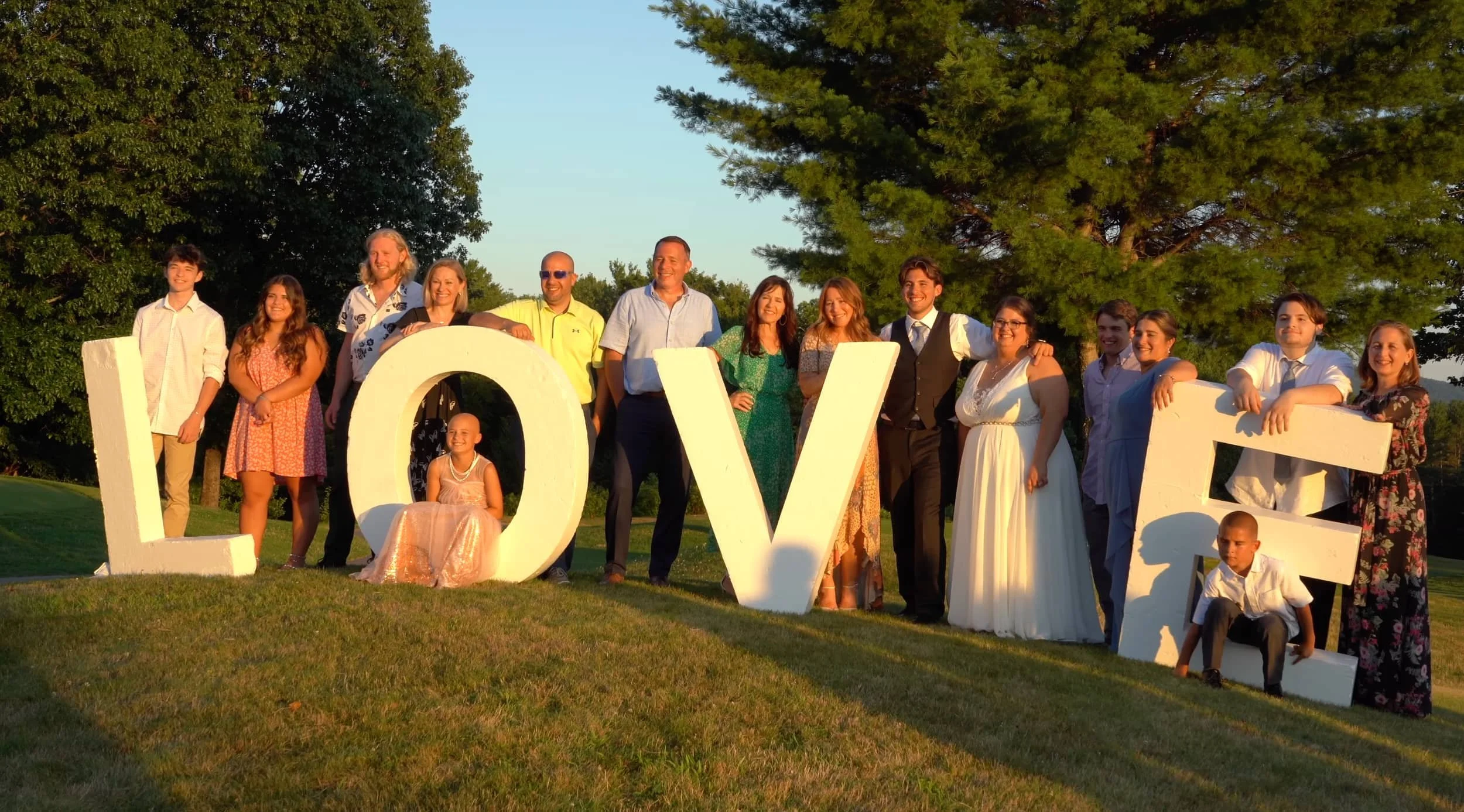 A group of people standing outdoors behind large white letters spelling "LOVE" on a grassy area with trees and a clear sky in the background during sunset.