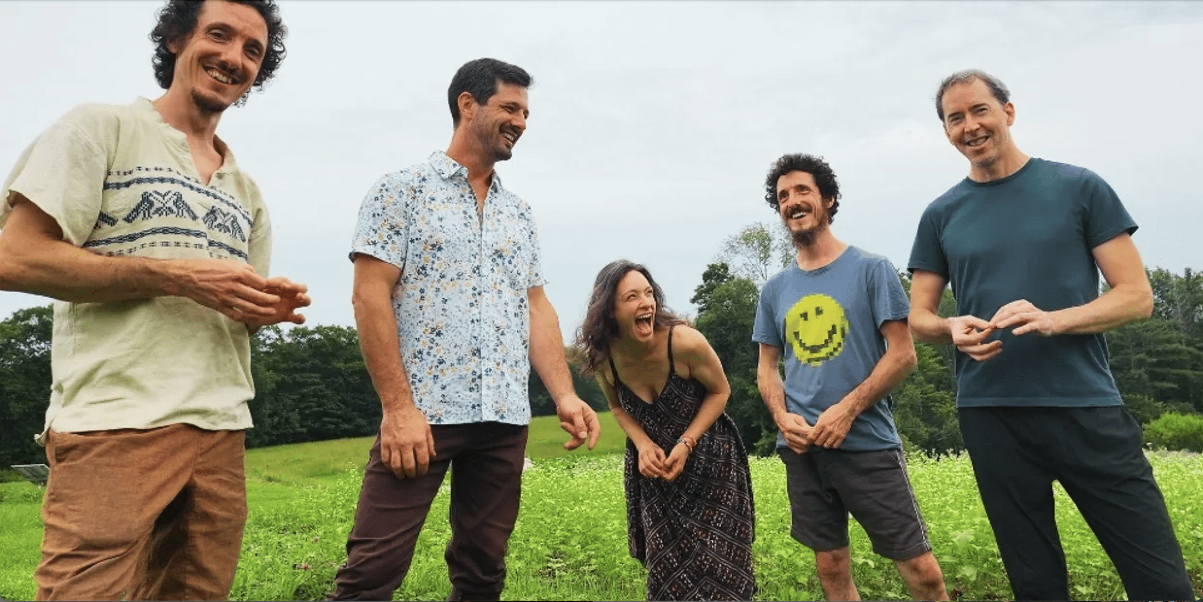 Five people standing outdoors in a grassy field, smiling and laughing.