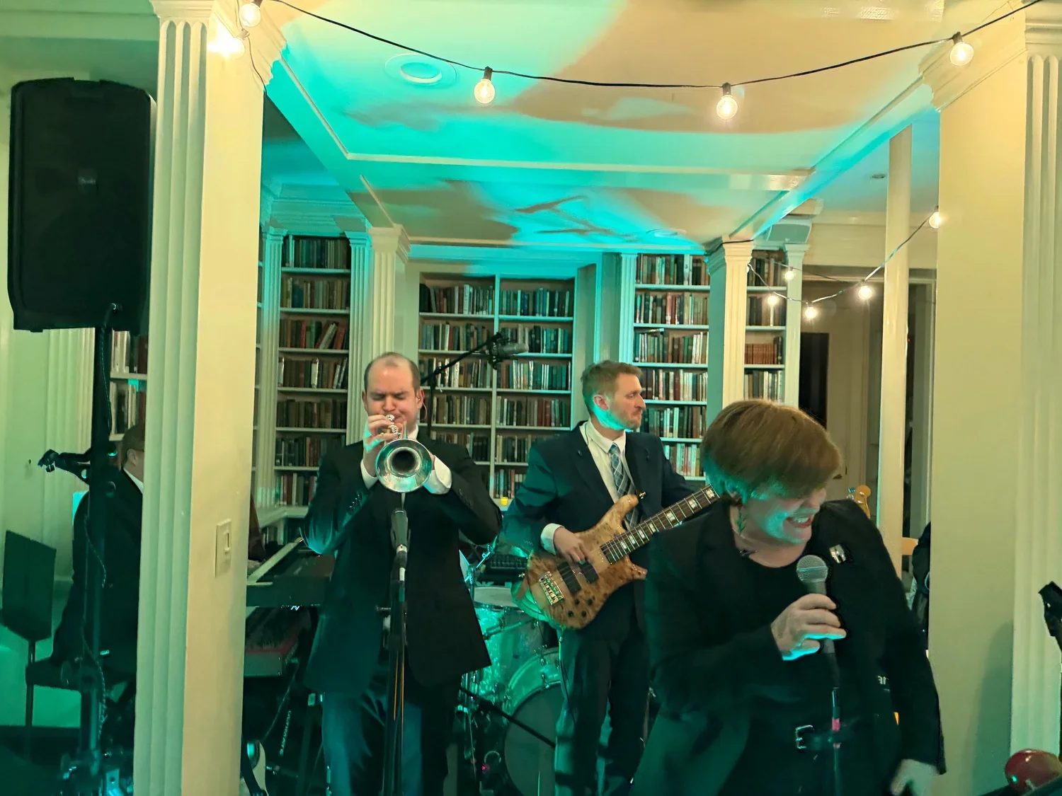 A band performing indoors with bookshelves and string lights, featuring a trumpet player, a bassist, and a woman singer holding a microphone.