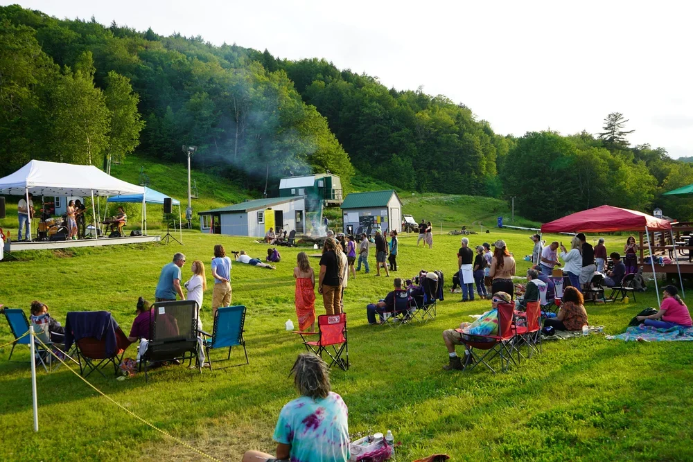 People gathered outdoors on a grassy hillside, some sitting in chairs and others standing, enjoying a community event with tents and a small stage, set against a backdrop of green trees and hills.