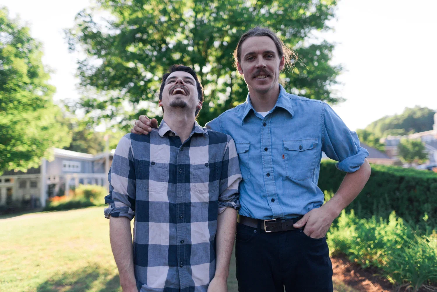 Two young men enjoying a walk outside on a sunny day, smiling and laughing, with green trees and houses in the background.