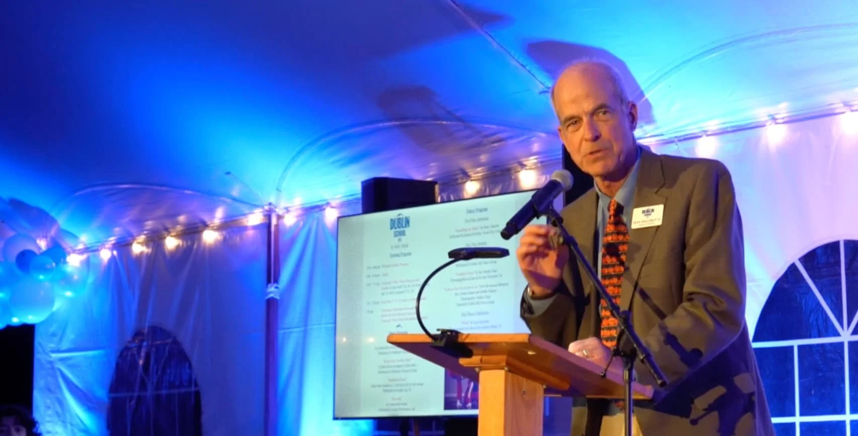 An older man in a suit and patterned tie speaking at a podium with a microphone, behind him is a screen displaying text, and the setting is illuminated with blue and purple lighting inside a tent with windows.