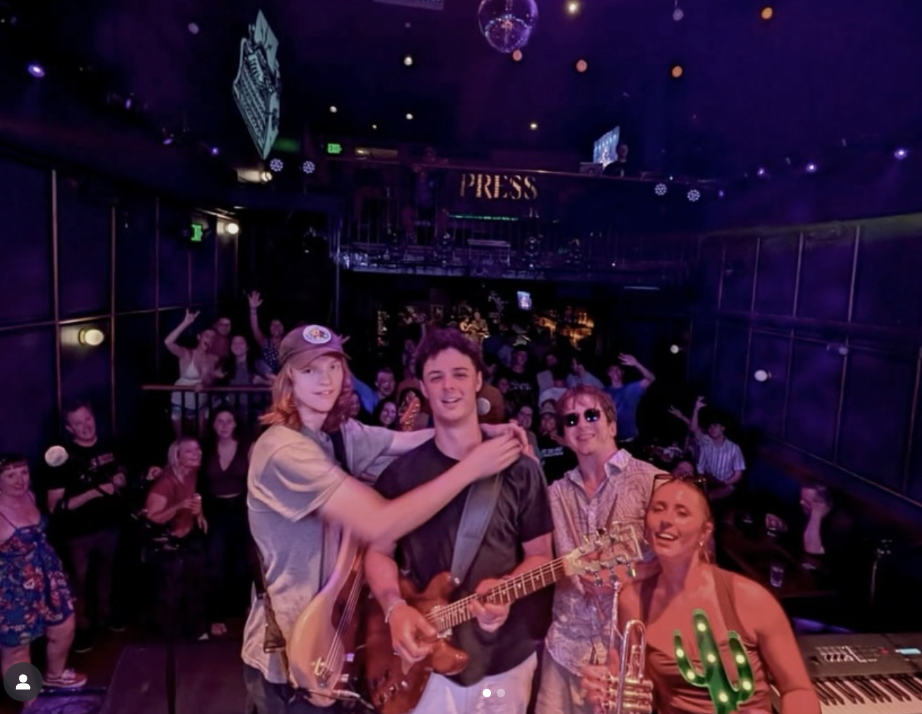 Group of people at a lively party or concert, with three individuals in the foreground holding musical instruments, smiling, and enjoying the moment, while others in the background dance and cheer in a dark, decorated venue with a sign that reads 'PRESS'.