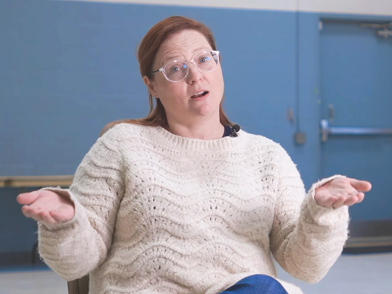 A woman with red hair and glasses wearing a cream-colored sweater, sitting and gesturing with her hands, against a blue wall background.
