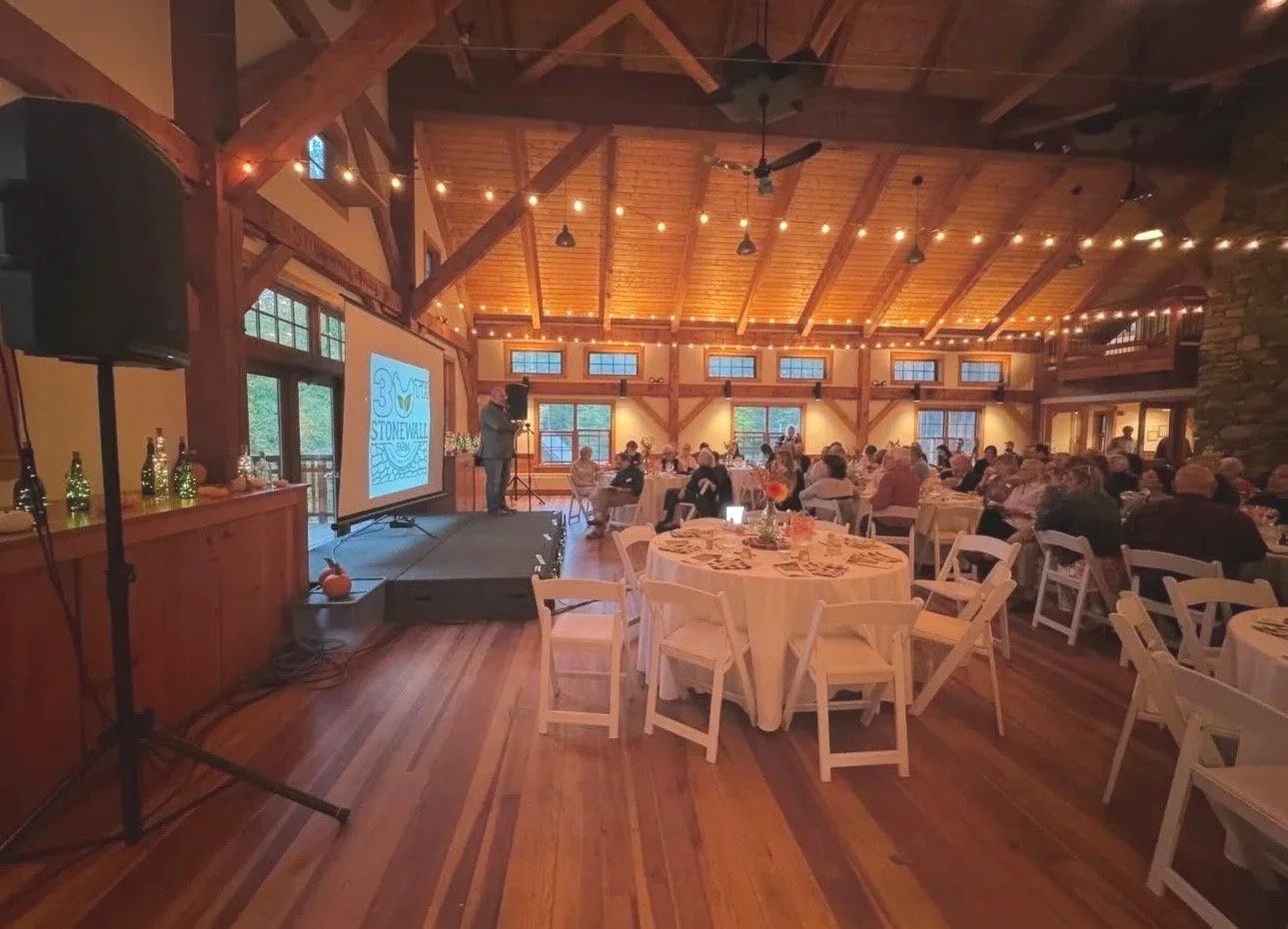 A spacious banquet hall with wooden beams and walls, decorated with string lights. Round tables with white tablecloths and chairs are set up for an event, with guests seated. A speaker stands on a small stage with a projector screen displaying the 30th anniversary logo of Stonewall. The room has large windows allowing natural light.