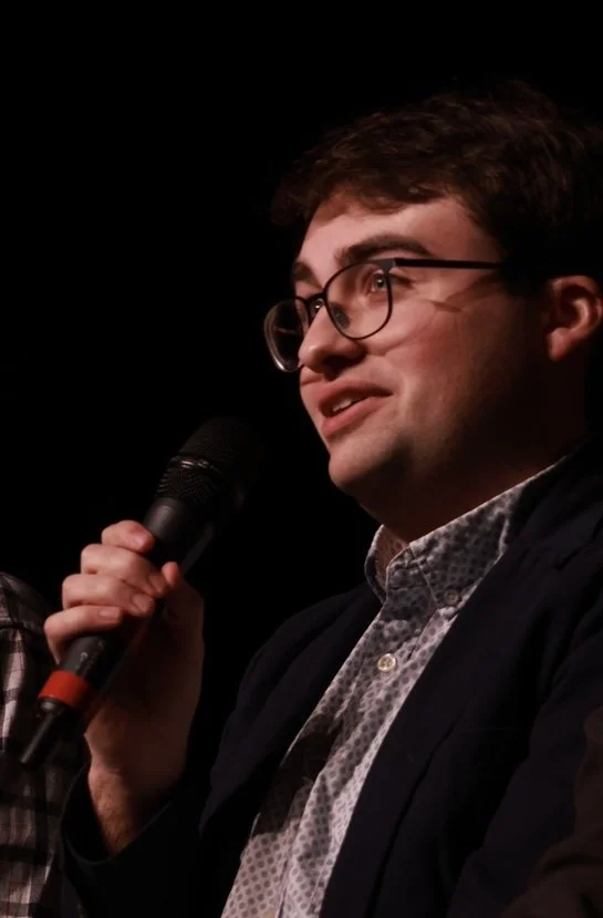 A young man with glasses holding a microphone, speaking on stage against a black background.