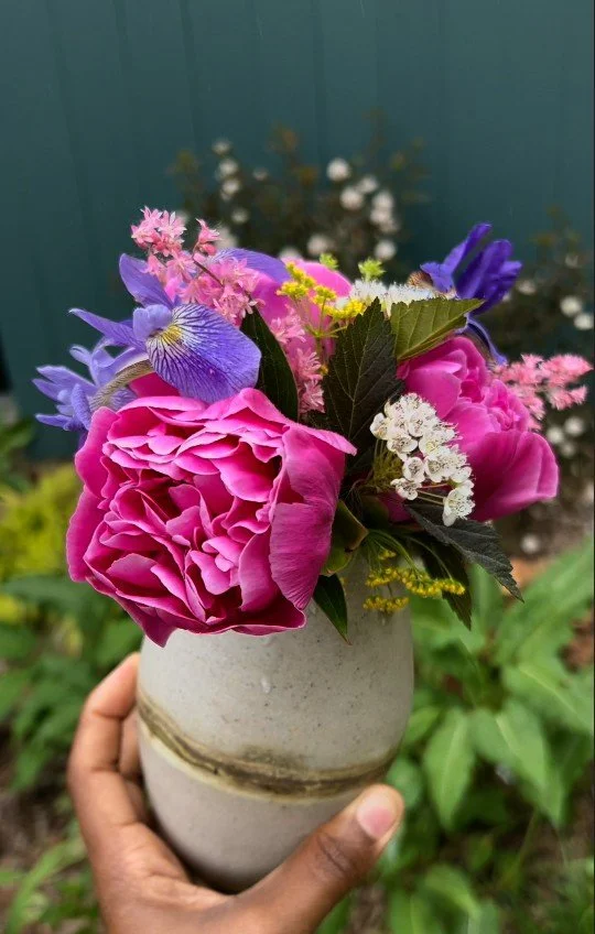 A hand holding a white ceramic vase with pink, purple, and white flowers against a green garden background.