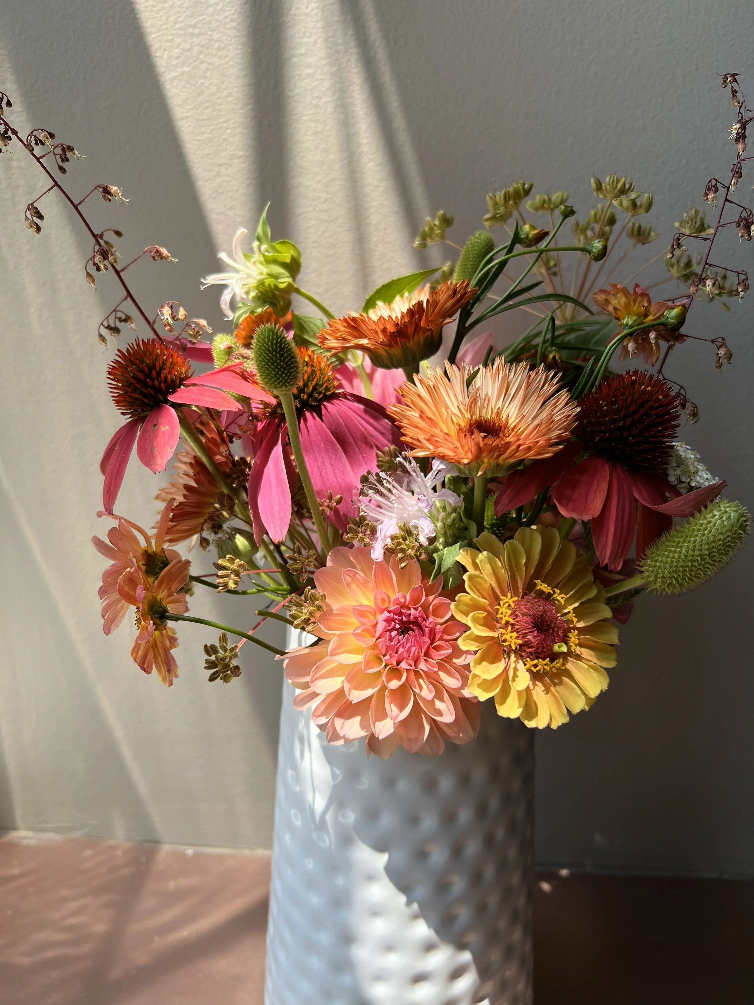 A bouquet of colorful flowers in a white textured vase, placed near a wall with sunlight casting shadows.