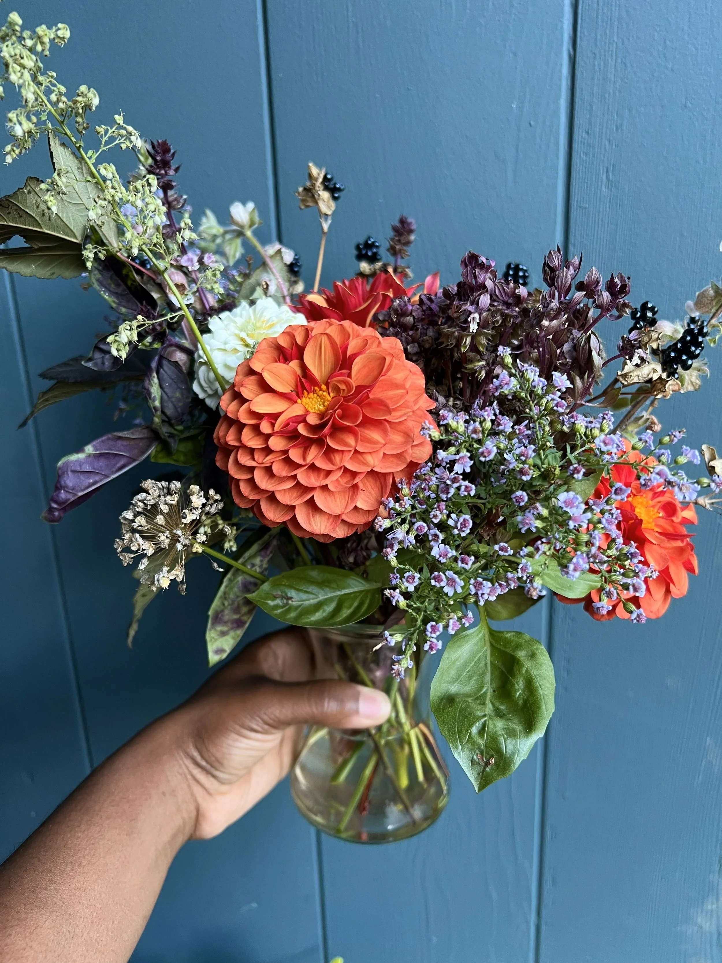 A hand holding a glass vase with a colorful bouquet of mixed flowers and foliage against a blue wall.