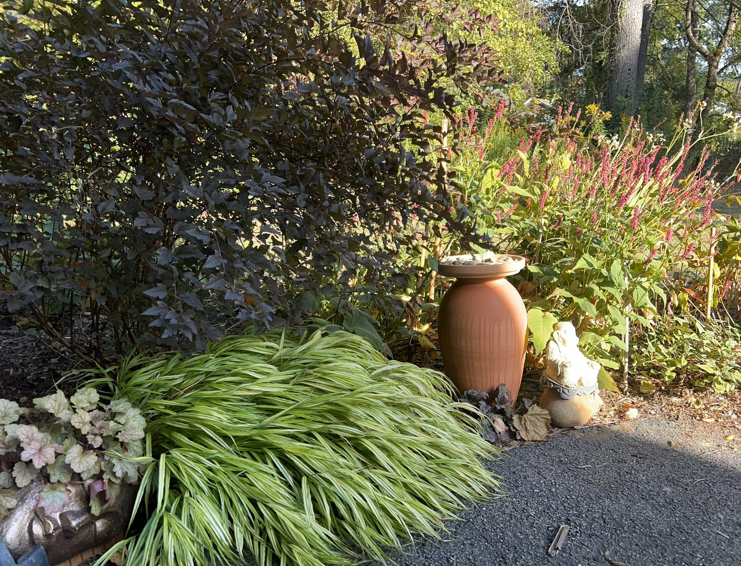 Garden scene with lush green plants, pink flowers, a large terracotta vase, and a small decorative lion statue.