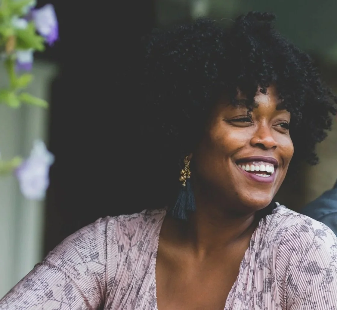 A woman with curly black hair smiling with eyes closed, wearing a patterned blouse and earrings, outdoors with blurred greenery in the background.