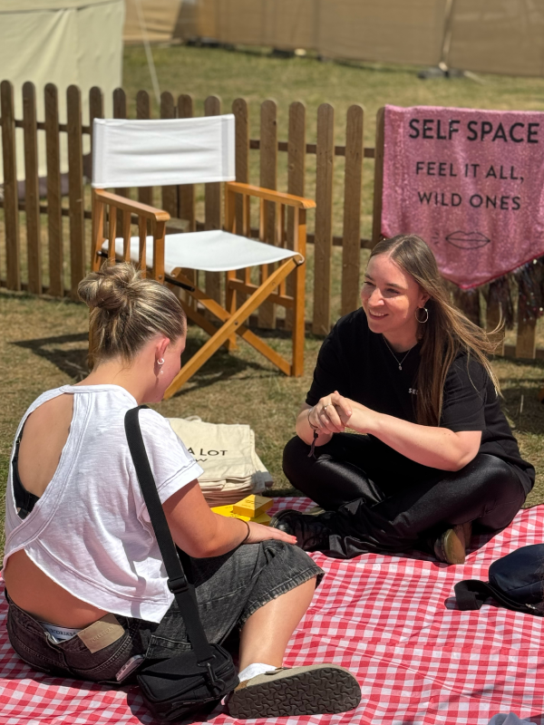Two women sitting on a red and white checkered blanket at outdoor event, engaging in conversation. Behind them is a wooden chair, a pink fabric sign with black text reading 'SELF SPACE FEEL IT ALL, WILD ONES,' and a wooden fence.