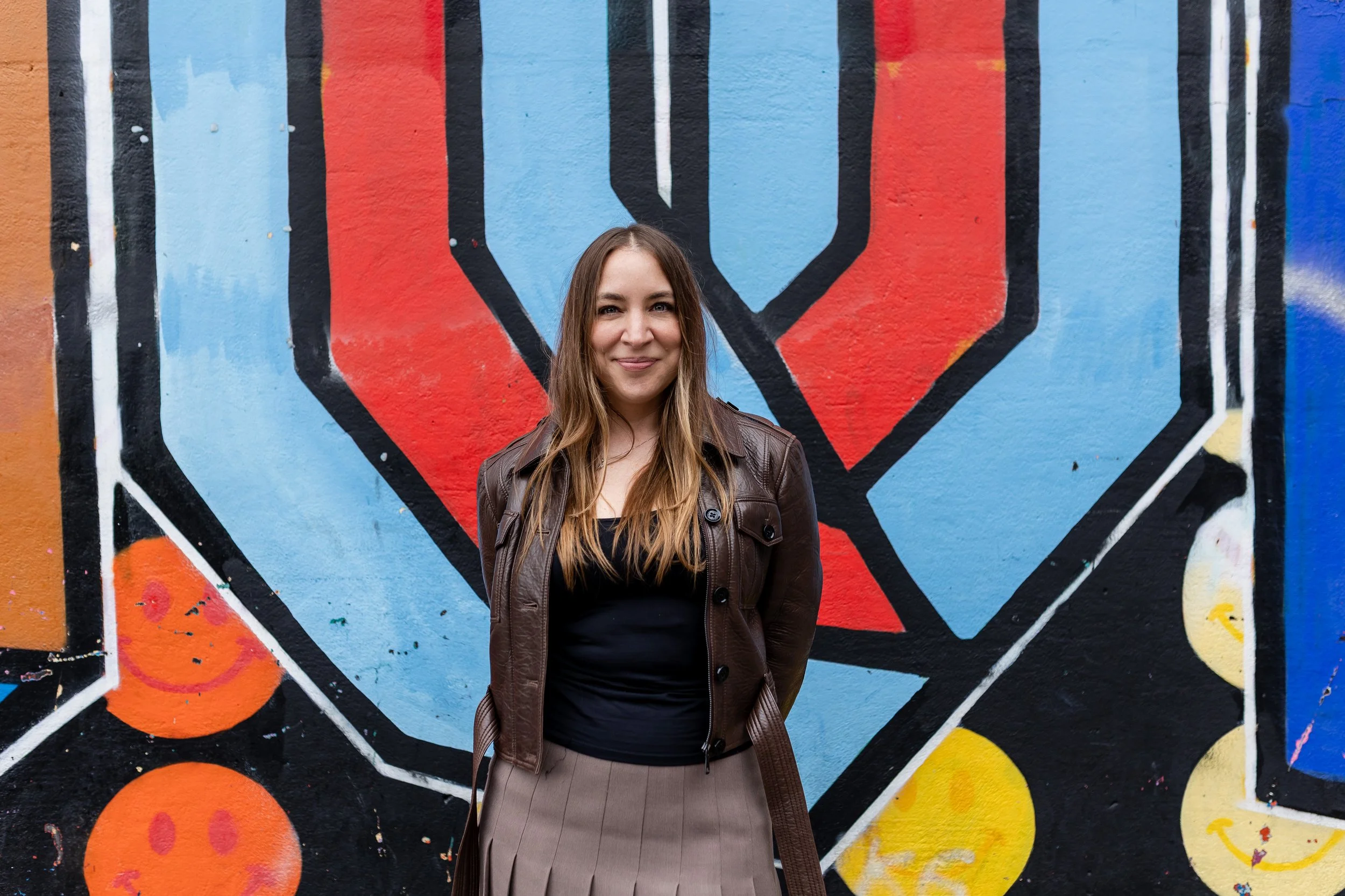 A woman with long brown hair smiling, wearing a brown leather jacket and black top, standing in front of a colorful graffiti mural with blue, red, orange, yellow, and black colors.