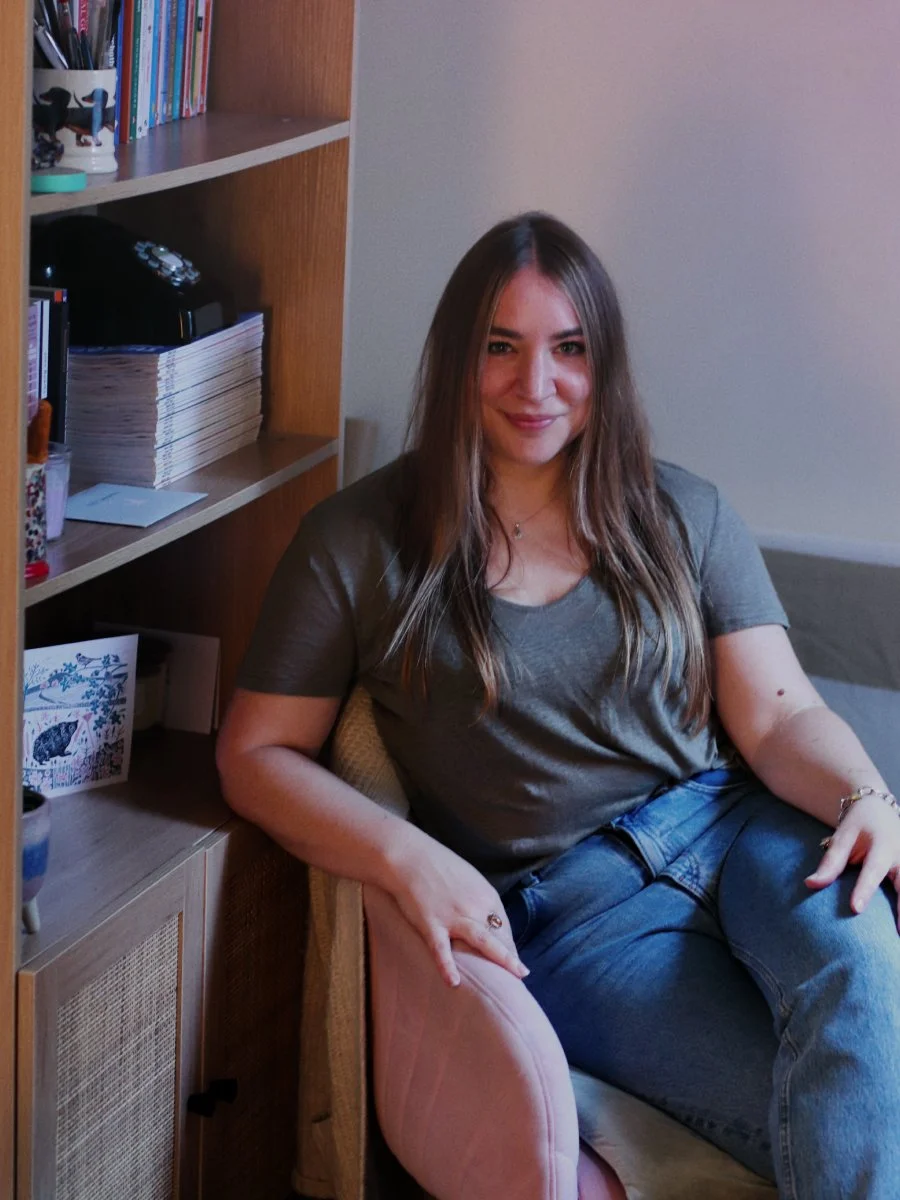 A woman with long brown hair sitting on a pink chair in a room next to a wooden bookshelf with books and magazines.