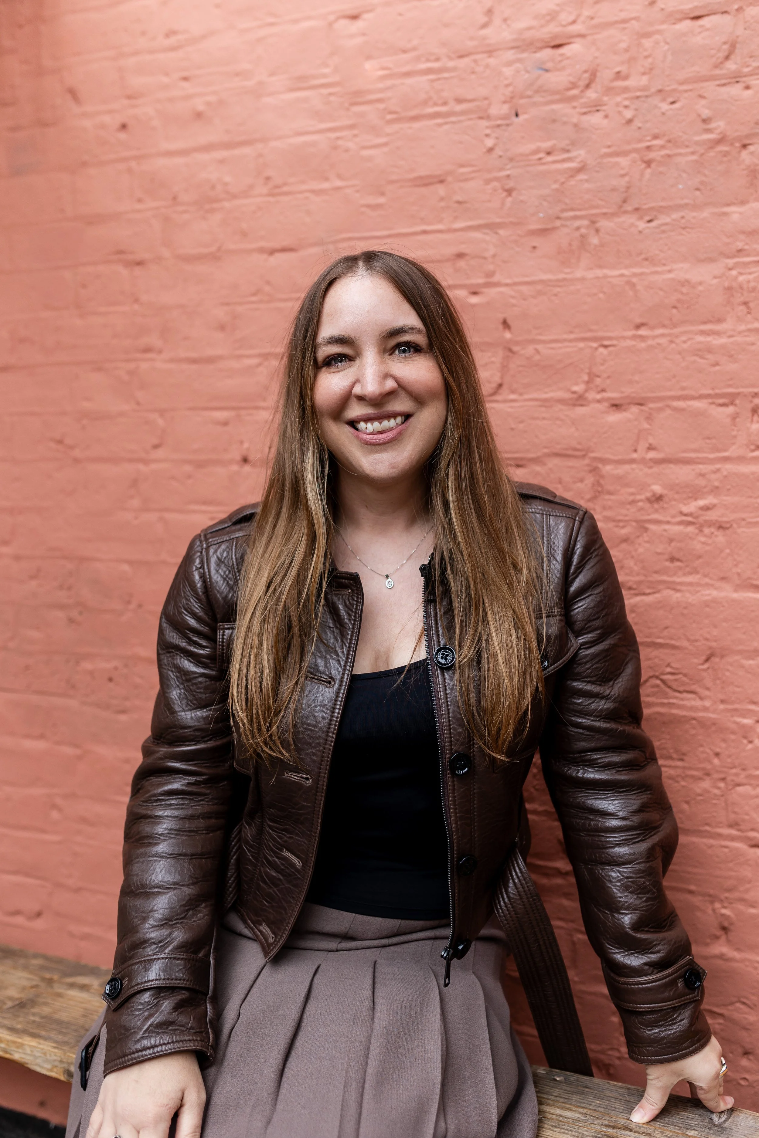 A smiling young woman with long brown hair, wearing a brown leather jacket over a black top, sits on a wooden bench against a pink brick wall.